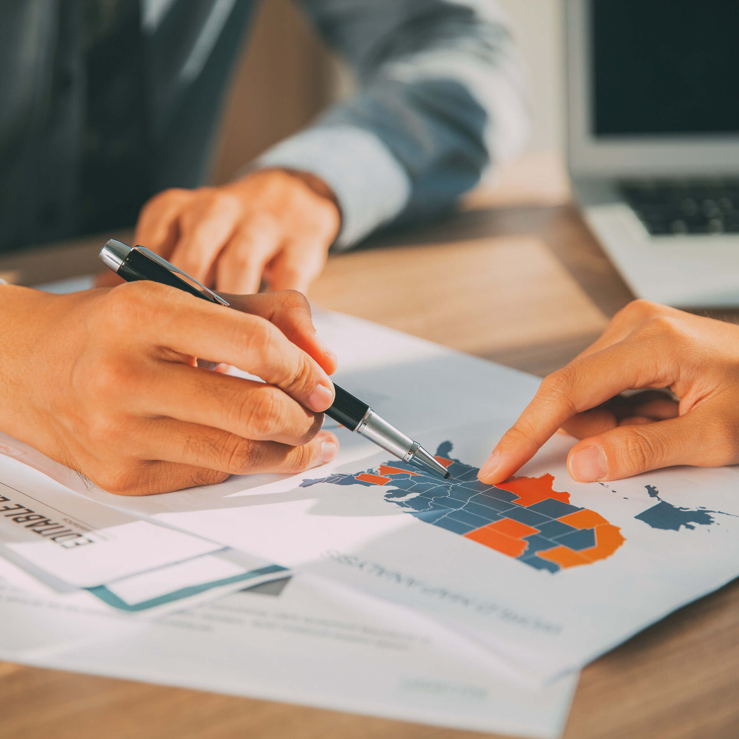 Two people discuss a printed map of the United States with regions highlighted in blue and orange. One person holds a pen and gestures at the map, while the other points to a specific state. A laptop is in the background.