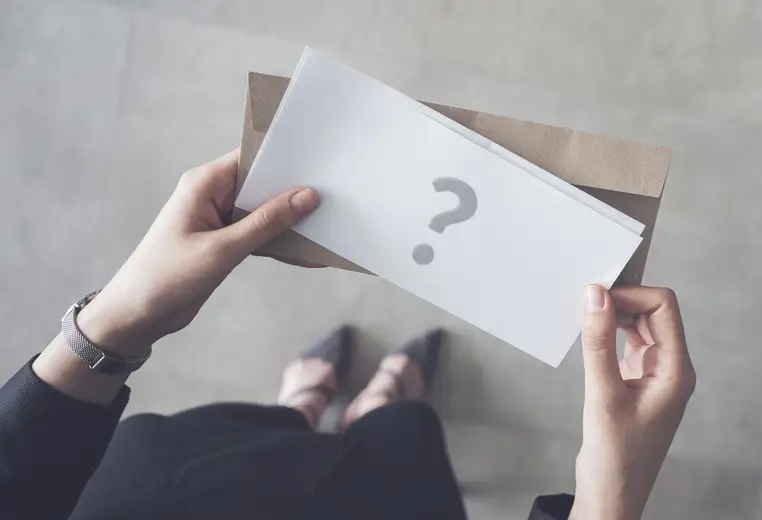 A person holds an envelope with a large question mark on the front. The background is a neutral gray surface, and the person is dressed in business attire.