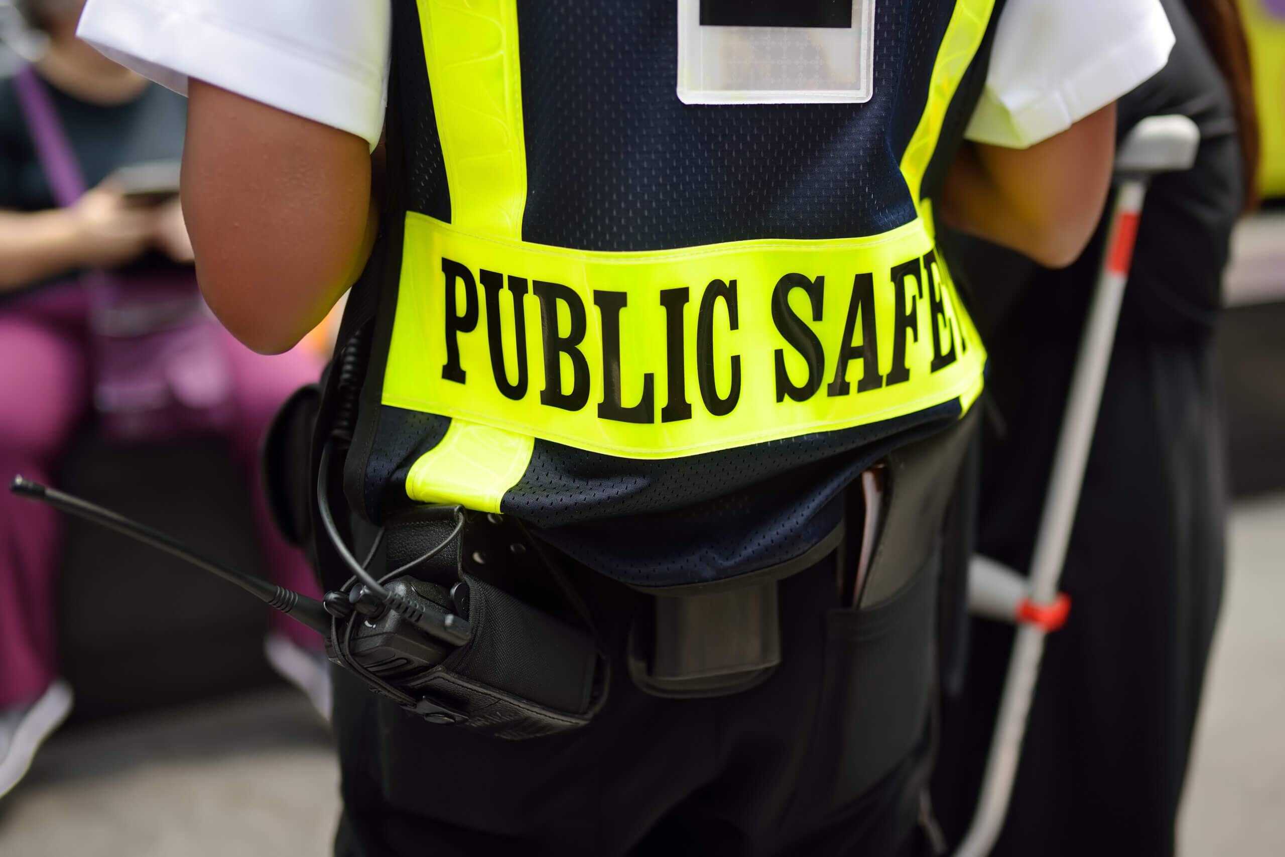 A person wearing a high-visibility vest with "Public Safety" written on the back is seen from behind. They have a radio attached to their vest. Others are partially visible in the background.