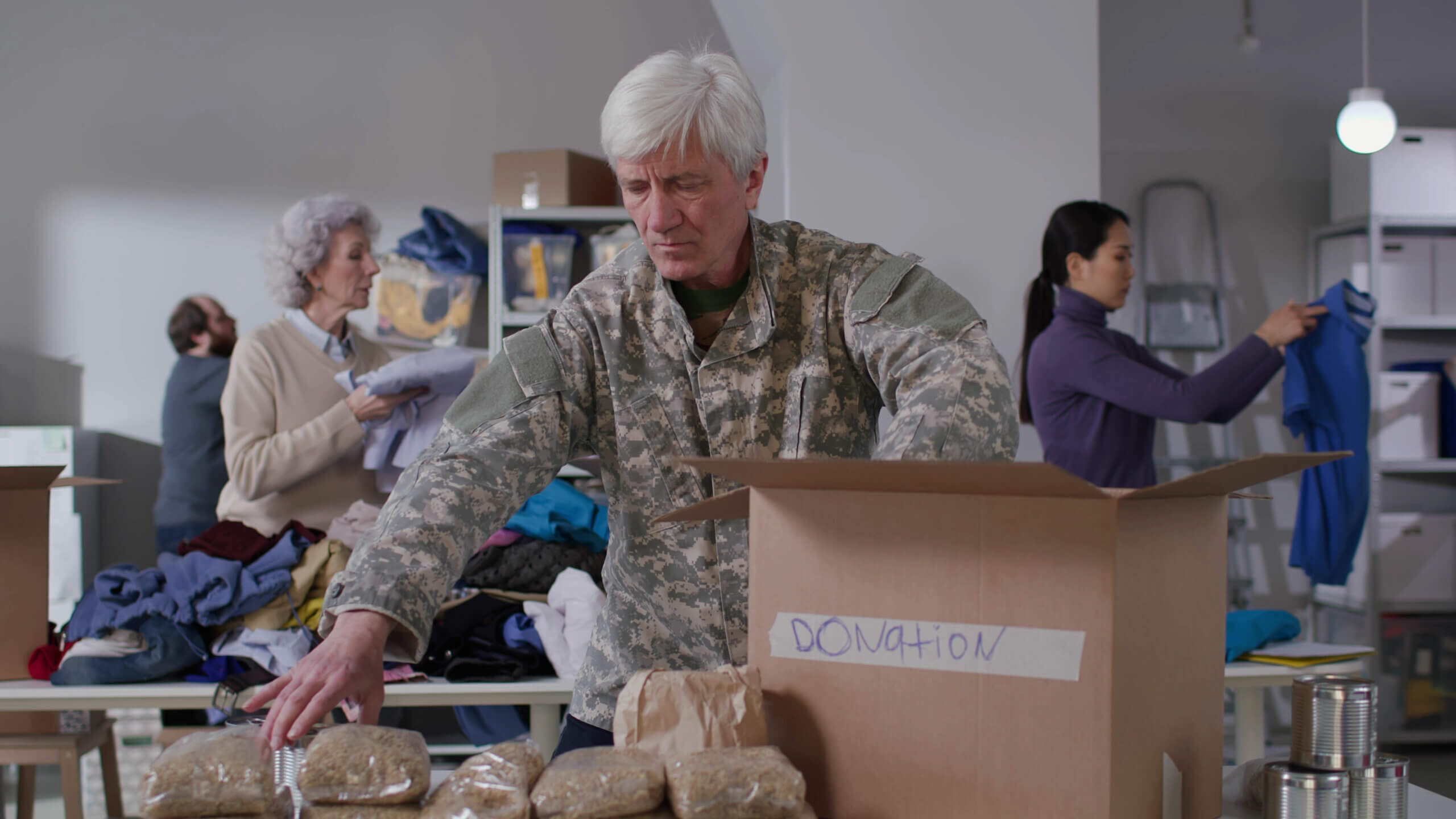 A man in camouflage uniform packs food into a "donation" box. In the background, an older woman organizes clothes and a younger woman folds garments. The setting is a donation center with shelves of sorted items.