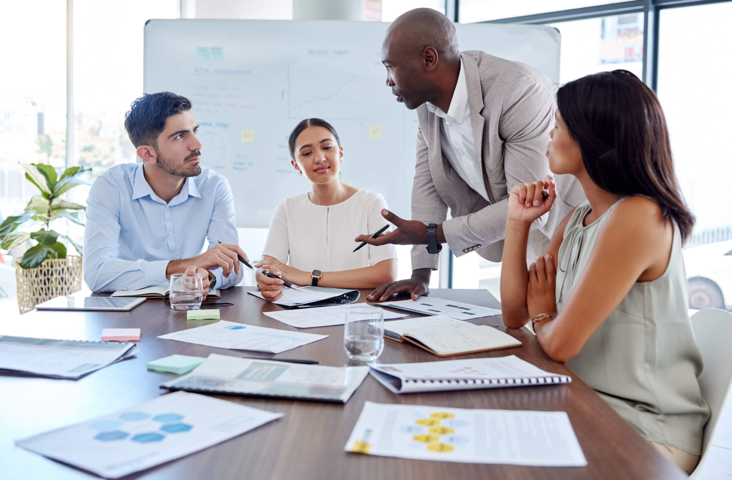 A diverse group of four people in a modern office setting are engaged in a collaborative discussion. They are seated at a table with papers and a laptop. A man in a suit is standing, actively speaking to the others. A whiteboard is in the background.