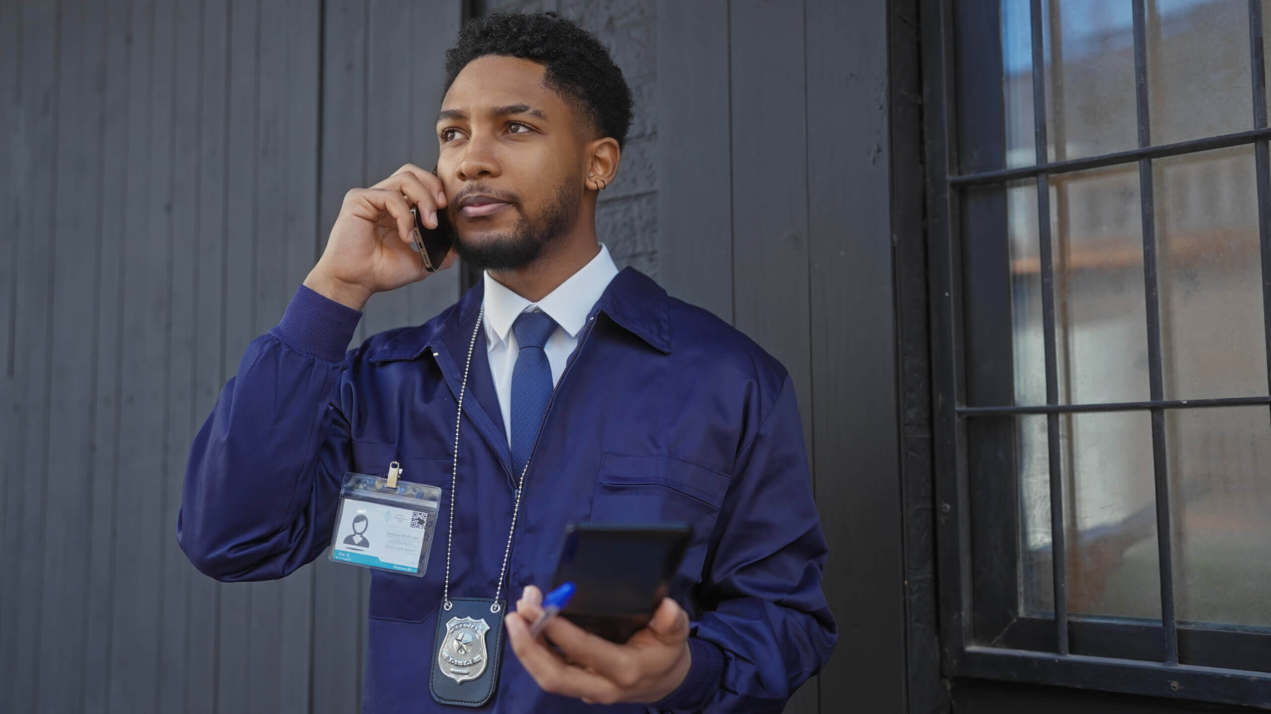 A man in a blue uniform is standing in front of a dark building, speaking on a cellphone. He holds a walkie-talkie in his other hand. He wears a lanyard and a badge.