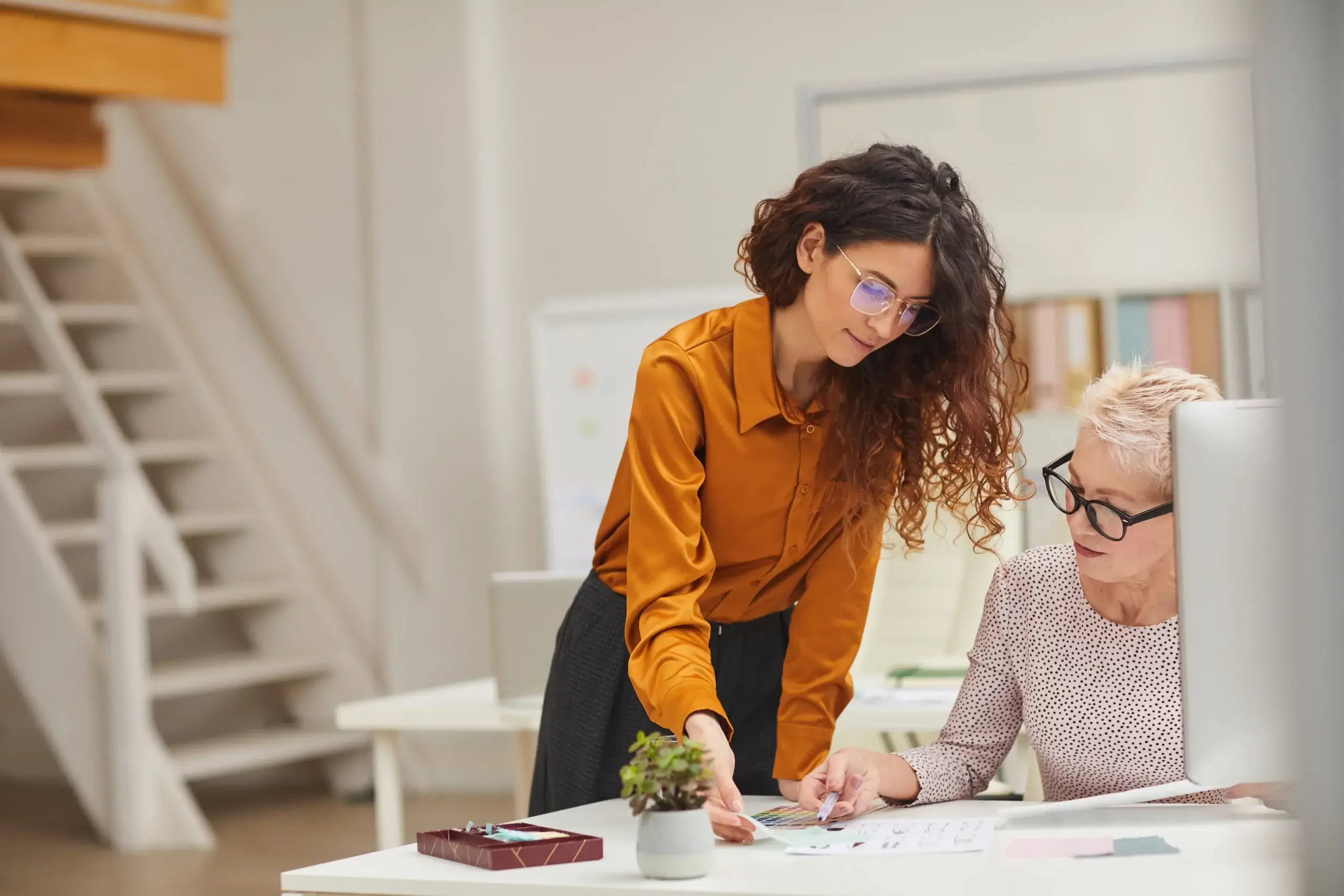 A woman with curly hair and glasses leans over a desk, discussing paperwork with an older woman who also wears glasses. They are in a modern office with shelves and a small plant on the desk.