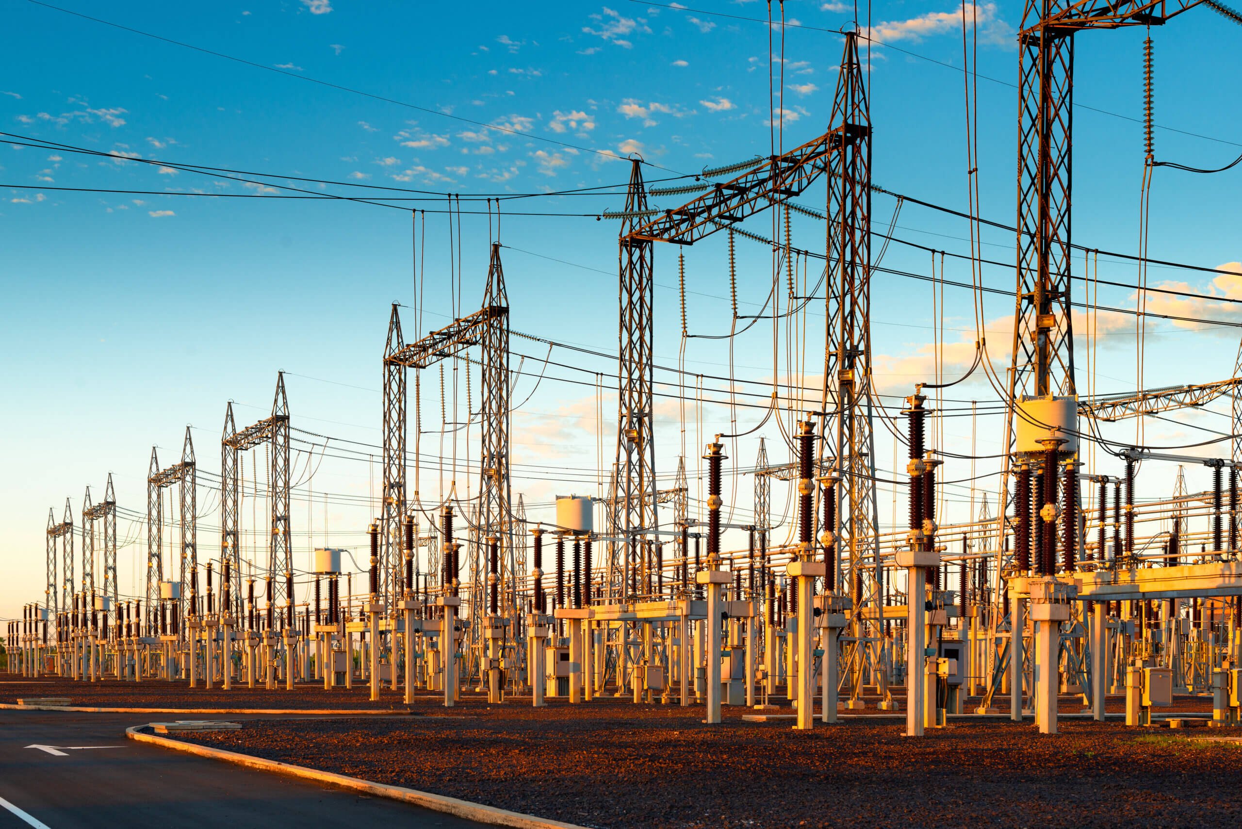 Power substation with numerous steel towers, electrical insulators, and wires against a blue sky at sunset. The ground is covered with gravel, and the setup is extensive, indicating a large-scale energy distribution facility.