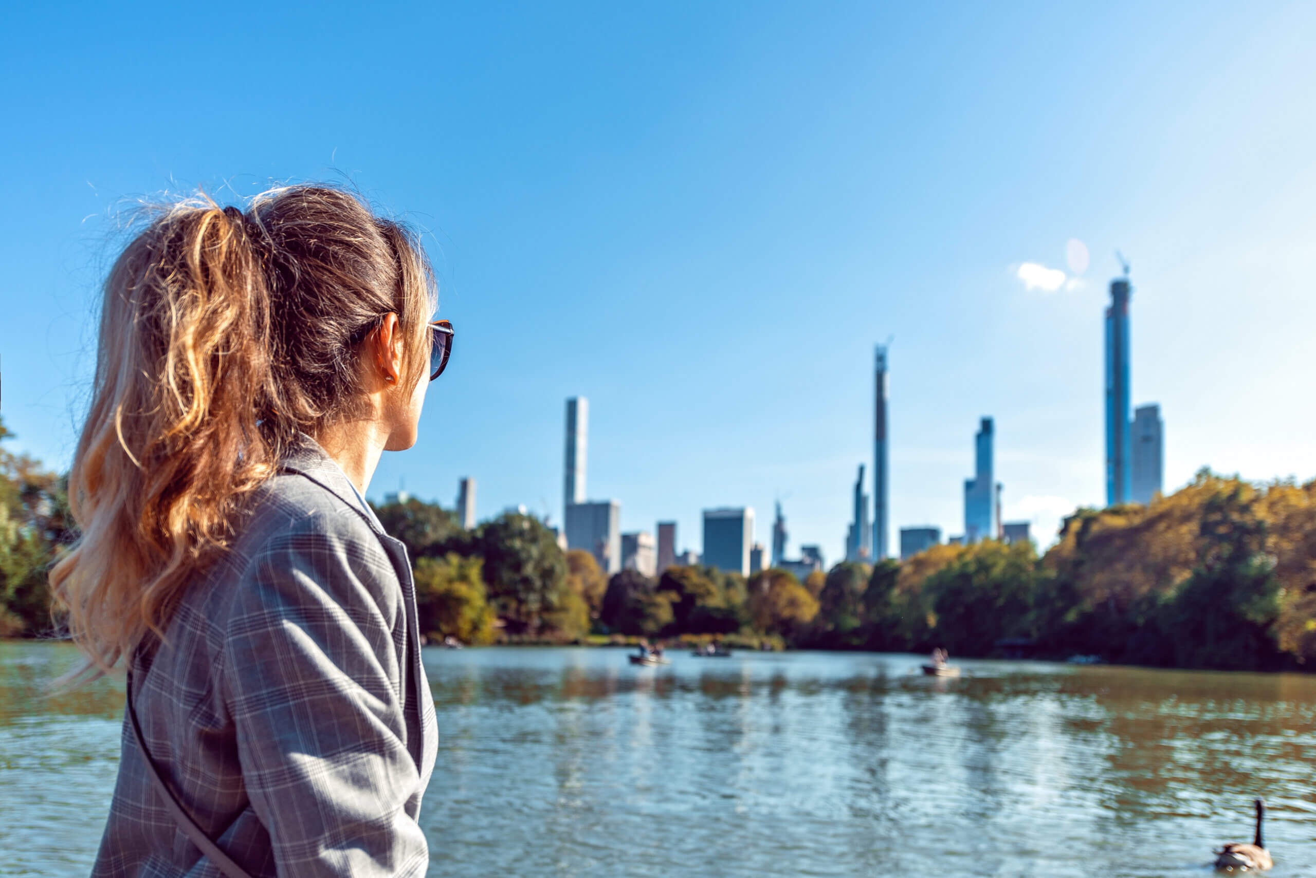 A woman with a ponytail and sunglasses stands by a lake, gazing at the New York skyline, where tall buildings rise under a clear blue sky. Trees line the lake's edge, whispering stories of nearby New York charities and their diligent audits by the state bureau.
