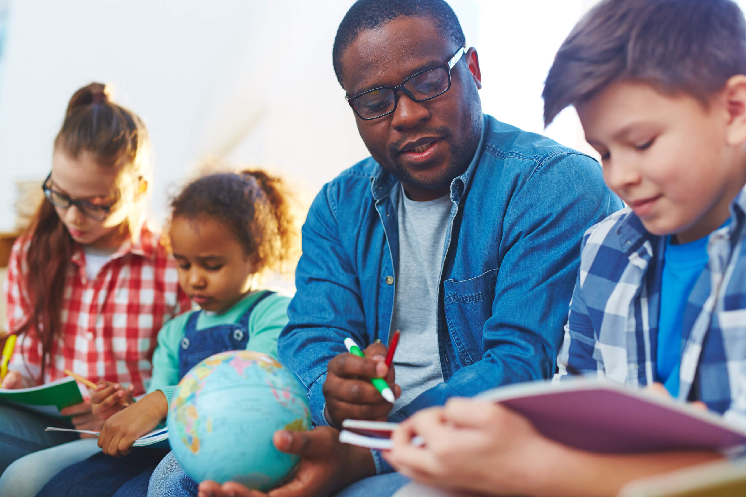 A teacher in glasses, wearing a denim shirt, is sitting with four children. One child holds a globe, while others are writing in notebooks. They are seated closely, suggesting a group activity or lesson. The setting appears educational and engaging.