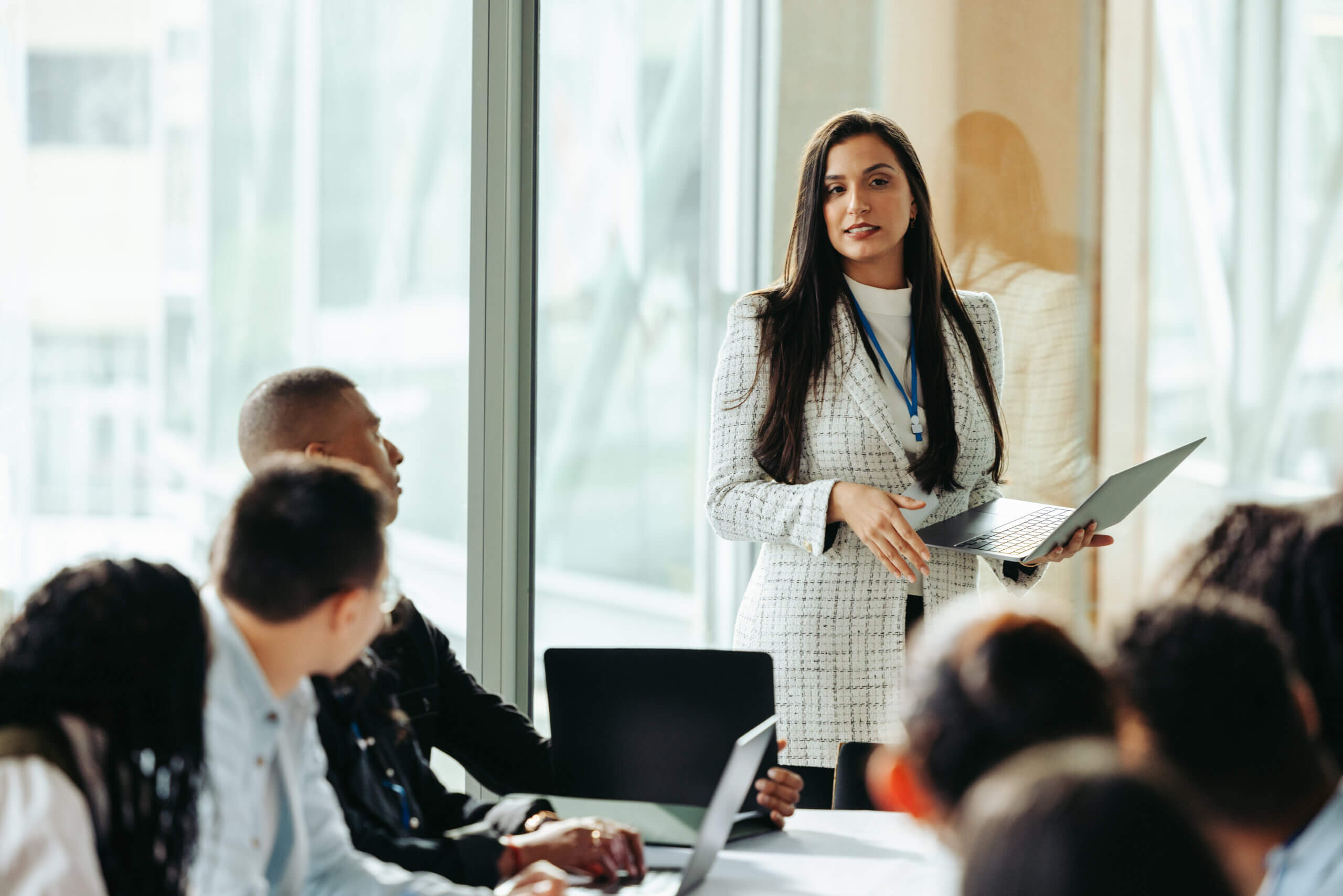 A woman in a white checkered blazer stands holding a laptop, speaking to a group of people seated around a table. The room has large windows, and the participants are focused on her presentation.