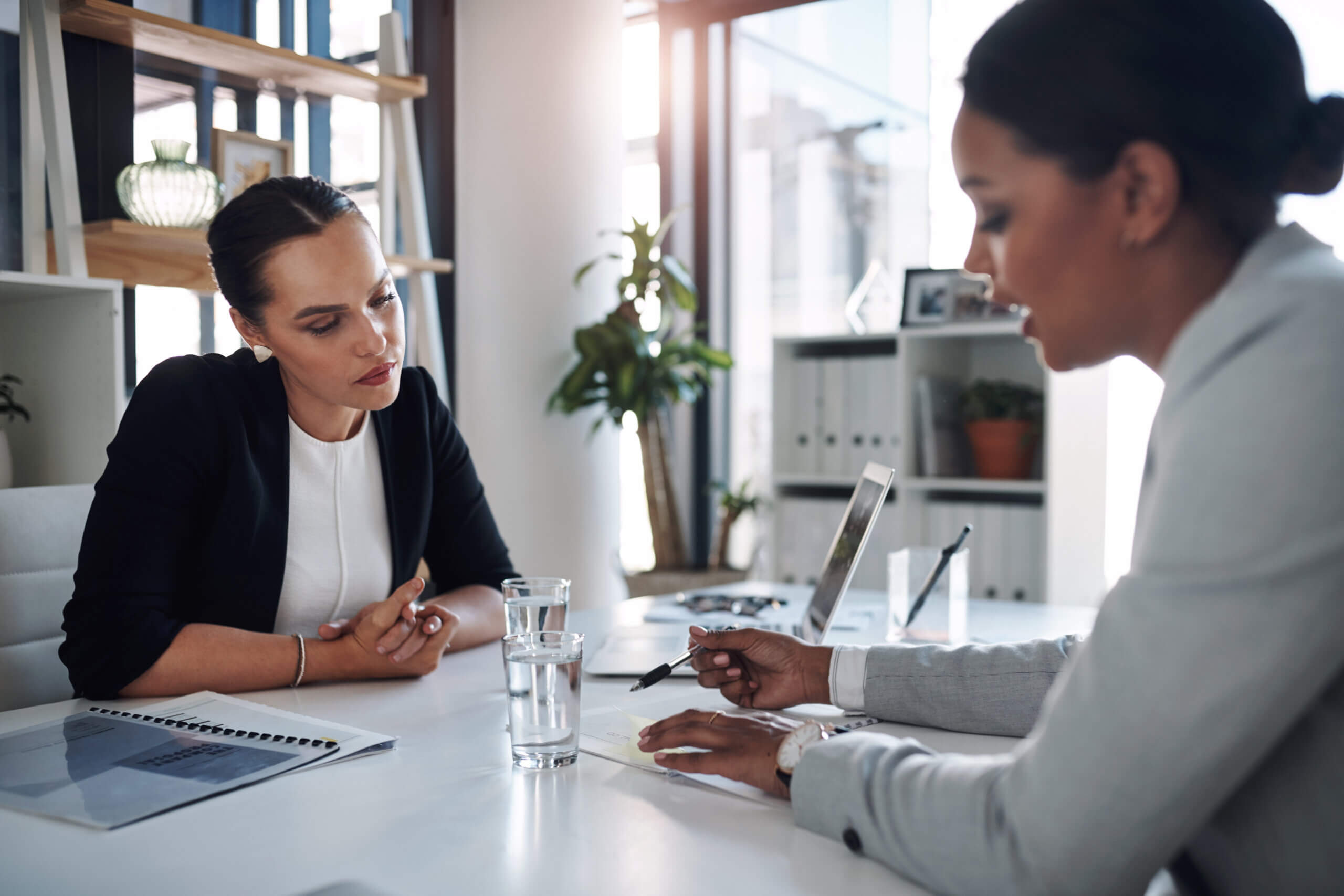Two women sitting at a desk in a modern office. One is explaining details, pointing with a pen, while the other listens attentively. A laptop and a glass of water are on the desk. Shelves and a plant are visible in the background.