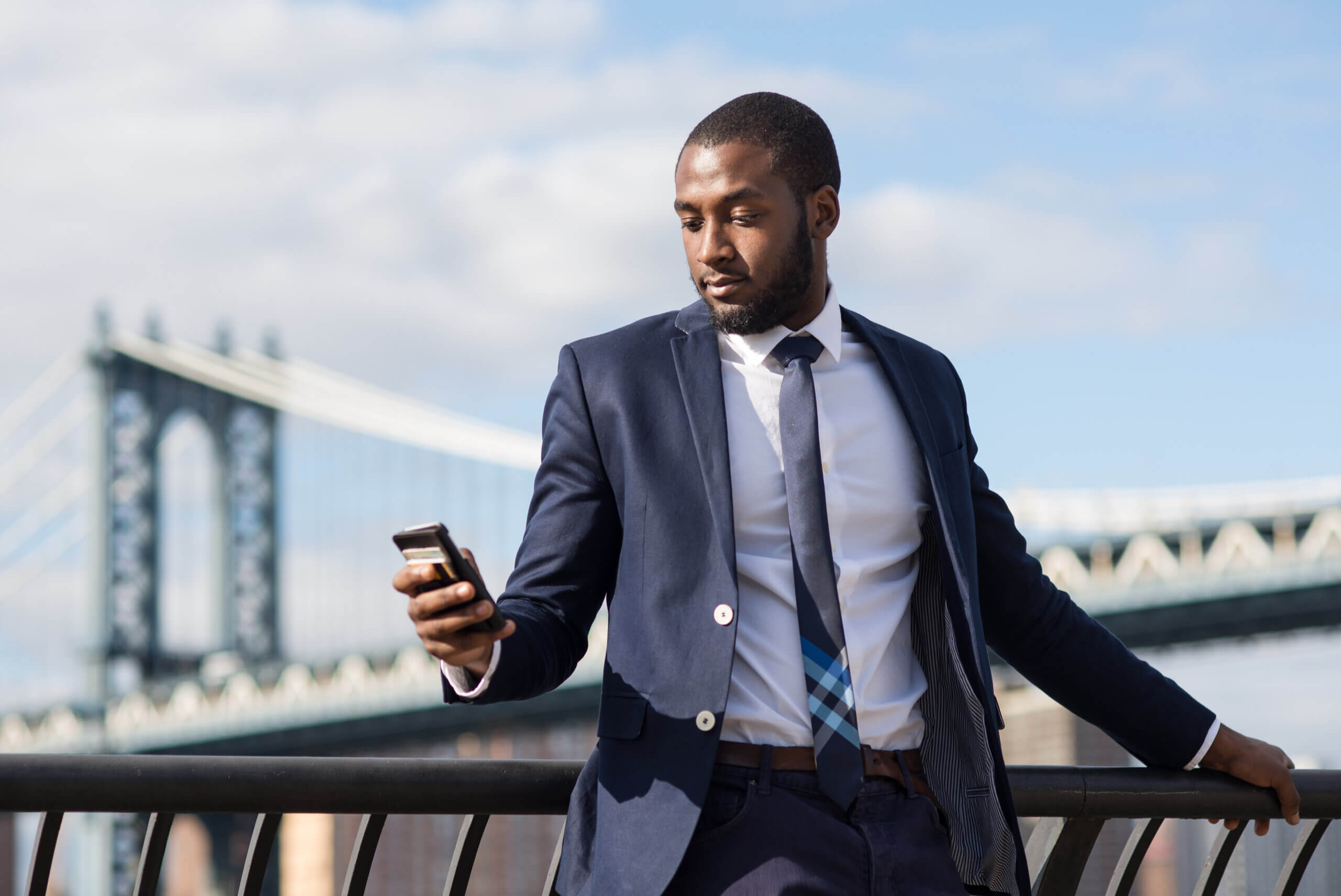 A man in a navy suit and striped tie looks at his phone while leaning against a railing. Behind him, a large suspension bridge stretches across a partly cloudy sky.