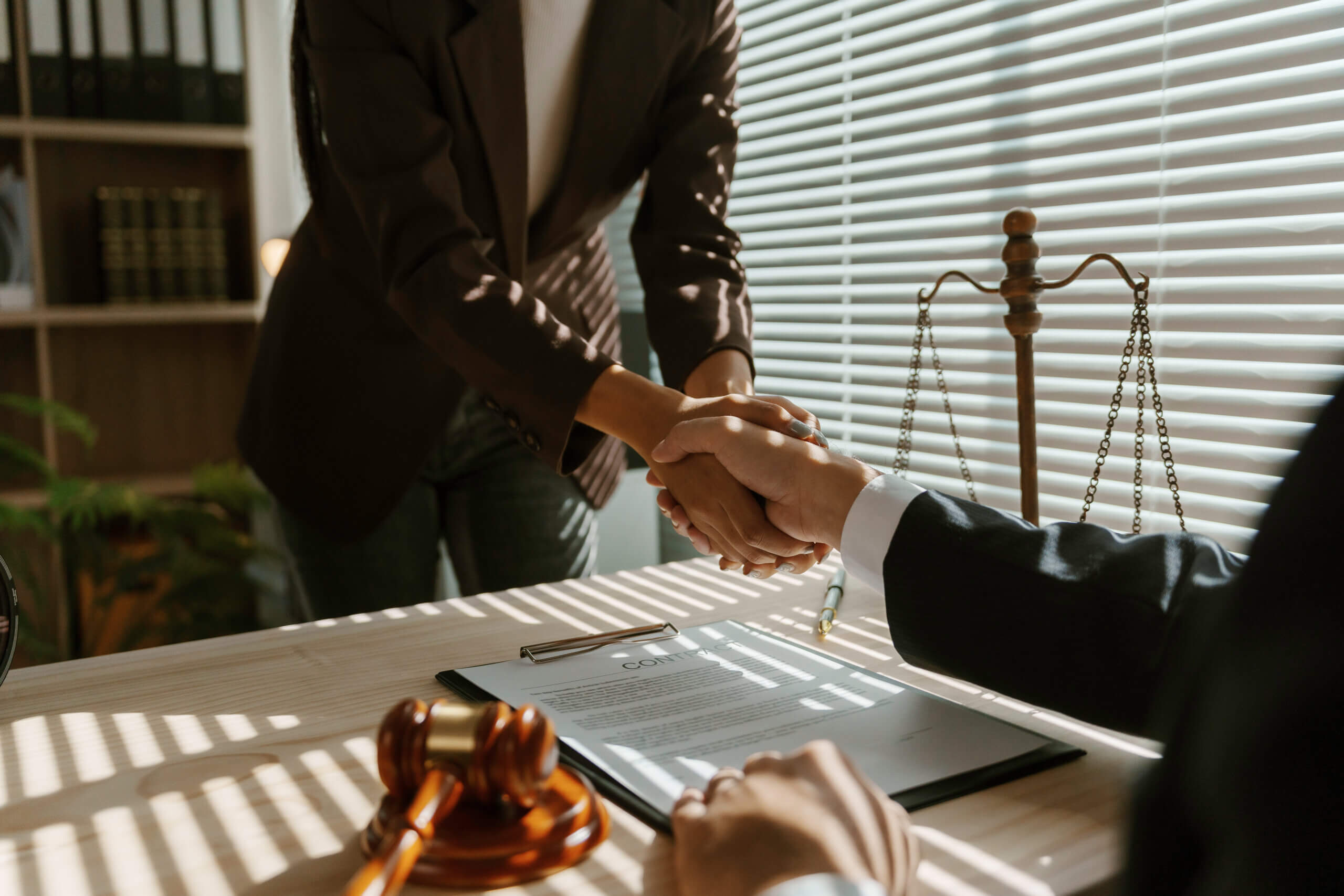 Two people are shaking hands across a desk in a bright office. The desk has documents, a gavel, and scales of justice. Sunlight filters through window blinds, creating striped shadows. Both are dressed in business attire.