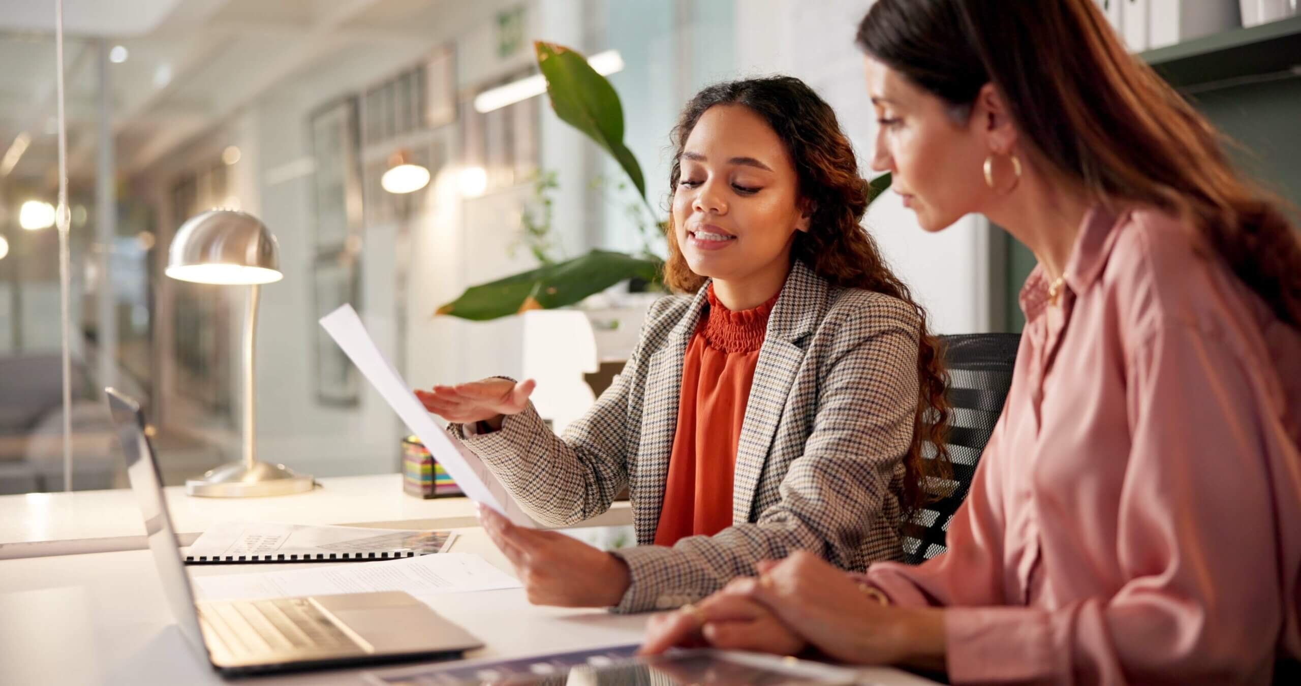 Two women sit at a desk in a modern office, reviewing documents. One woman, in a plaid blazer, gestures towards the paper, while the other in a pink blouse listens intently. A laptop, lamp, and plant are visible on the desk.