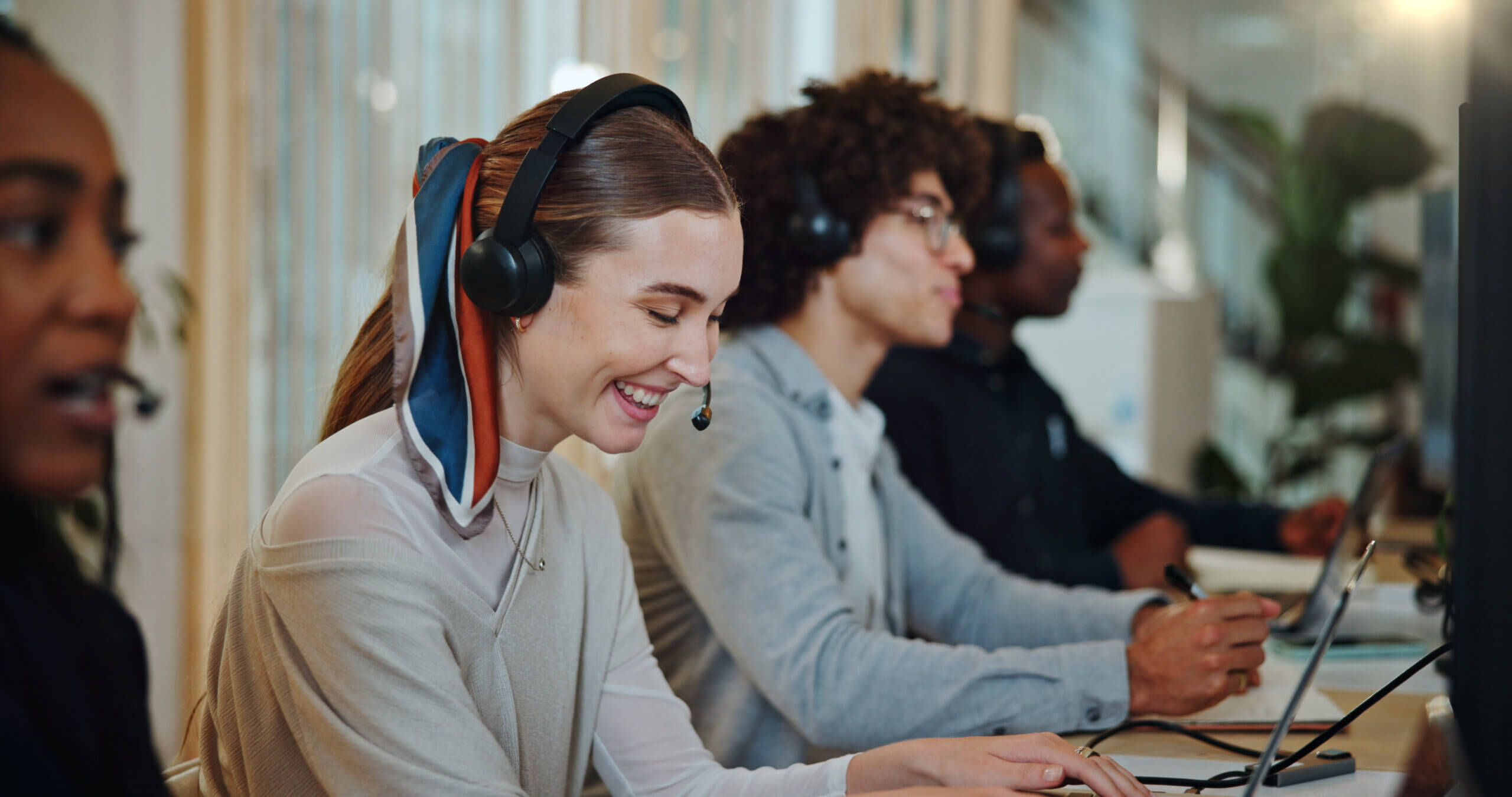 Four people sit at desks wearing headsets with microphones, focusing on their computers. A smiling woman in the foreground wears a scarf tied in her hair, with others working intently beside her. They appear to be in a modern office or call center.