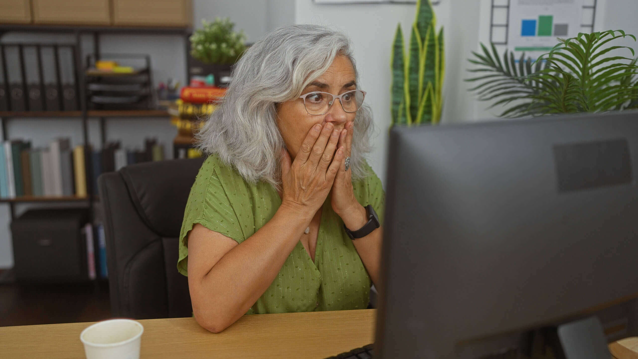 A woman with gray hair and glasses looks surprised while sitting at a desk in front of a computer. She is wearing a green shirt, with her hands covering her mouth, and is surrounded by office decor and plants.