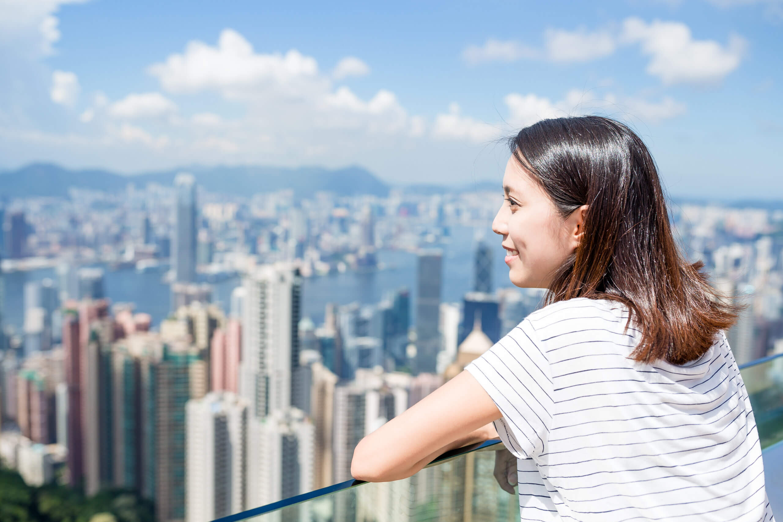 A woman with shoulder-length brown hair and a striped shirt is standing on a balcony, looking out over a cityscape filled with tall buildings and a distant harbor under a blue sky with scattered clouds.