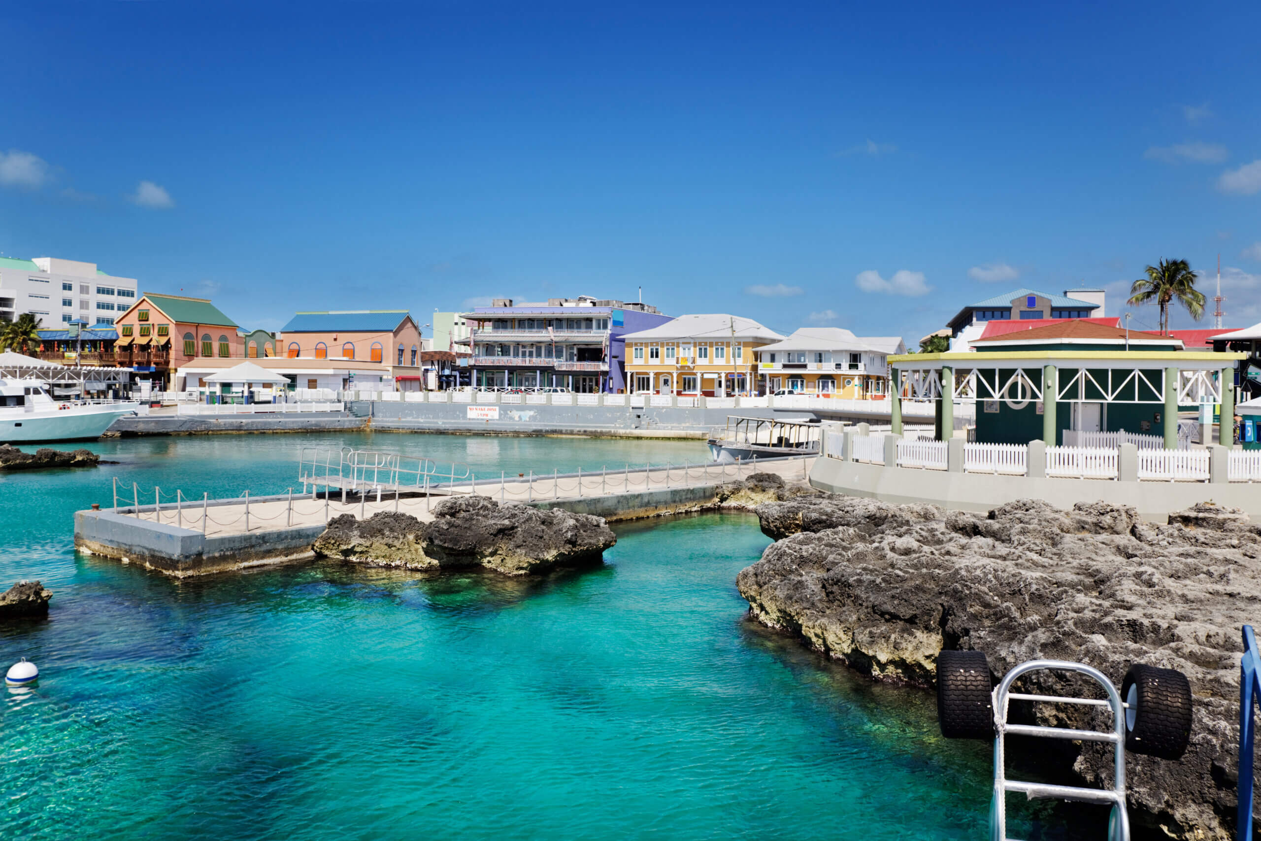 A vibrant waterfront scene with clear turquoise water and rocky shoreline. Colorful buildings line the coast under a bright blue sky, with a mix of palm trees and structures adding to the tropical atmosphere.