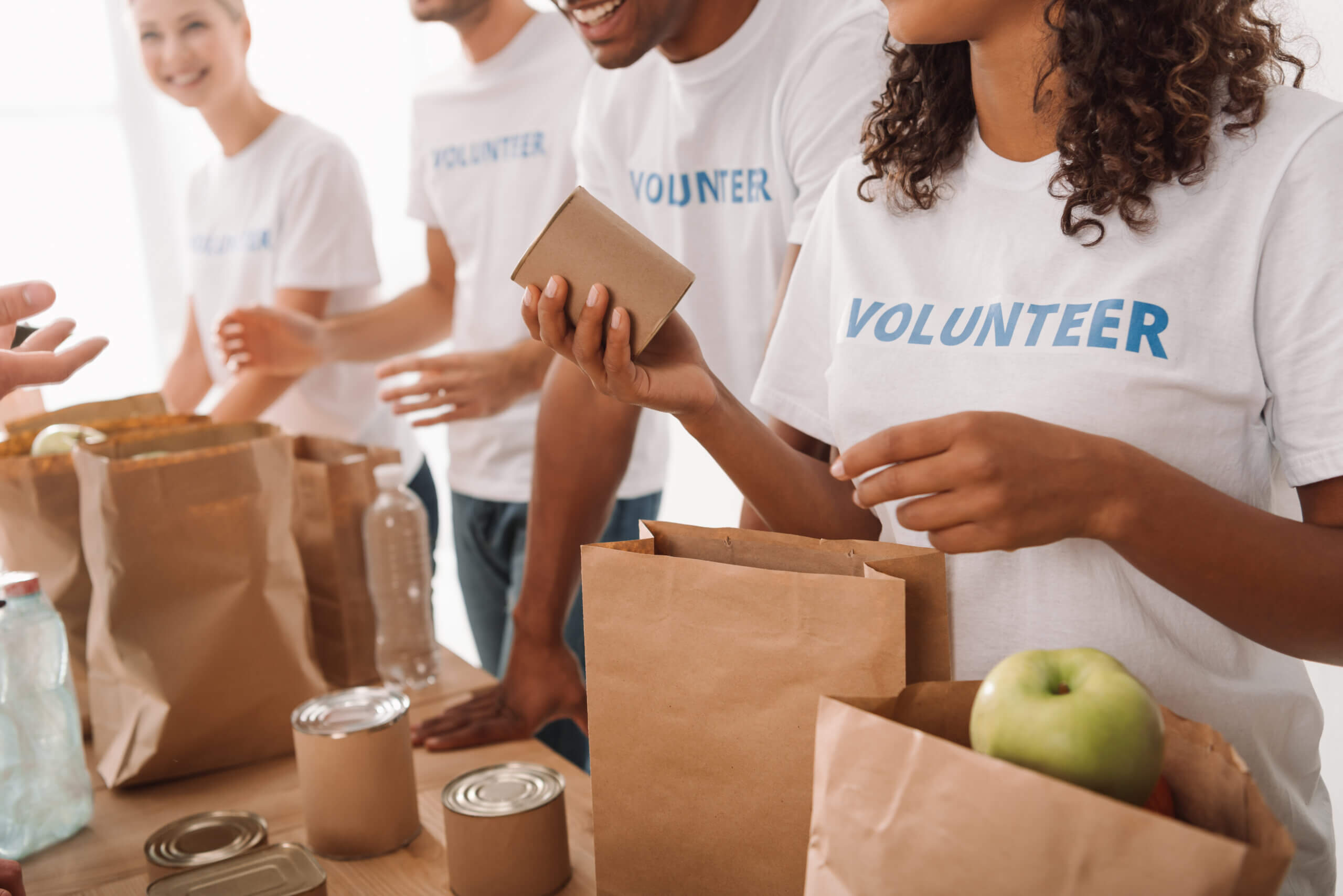 Volunteers in white t-shirts are organizing food donations at a table. They are filling paper bags with items like canned goods and a green apple. The word "Volunteer" is printed on their shirts.