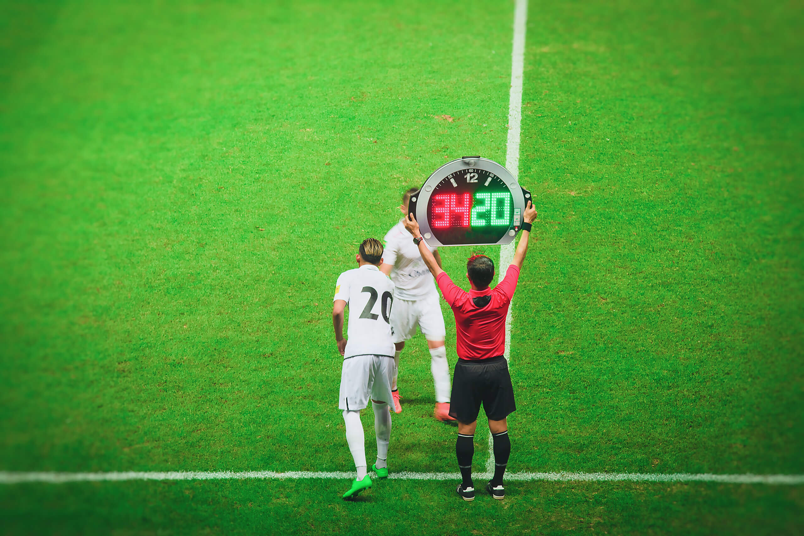 A soccer player in a white jersey, number 20, enters the field as a substitute, replacing another player. The referee holds up a digital substitution board showing the numbers 34 and 20. The lush green field is visible in the background.