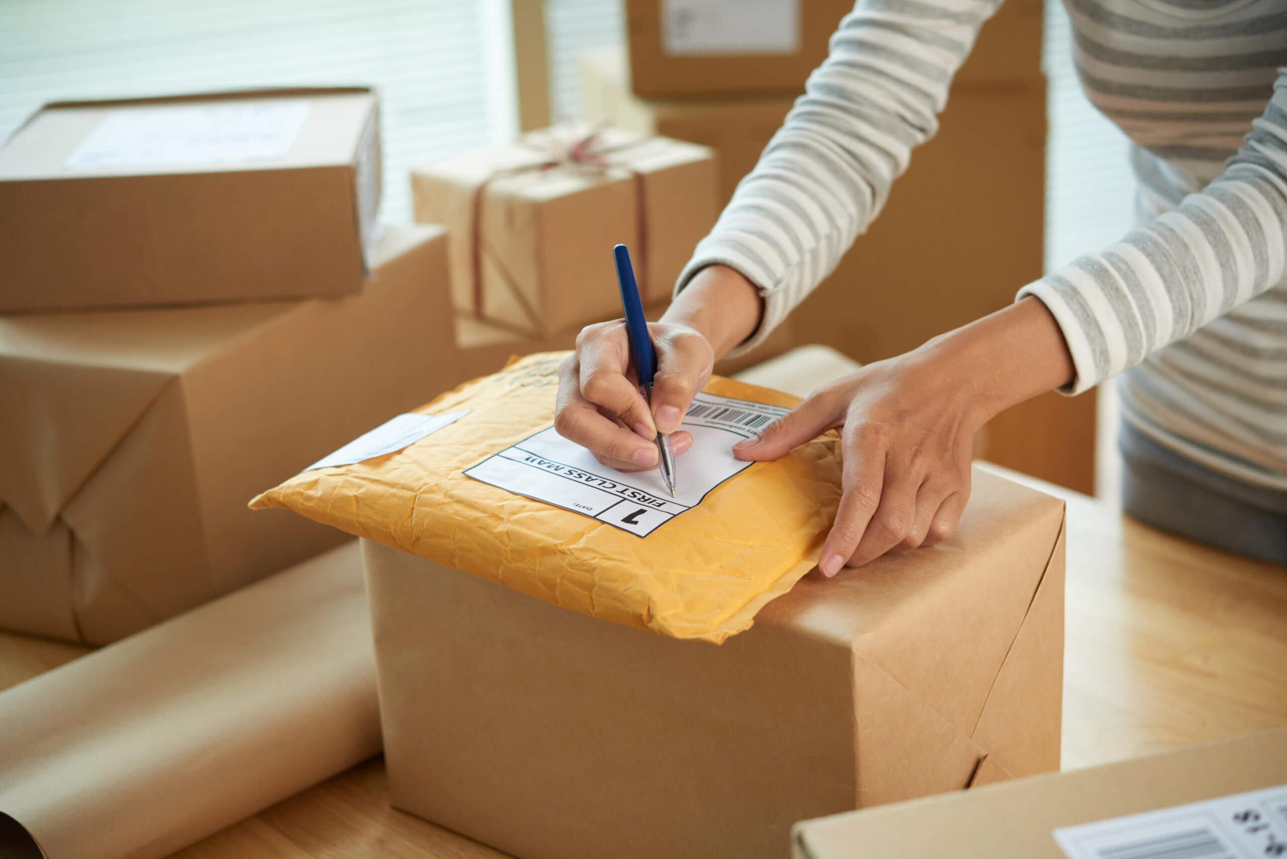 Person writing on a padded envelope with a pen. The envelope is placed on top of a cardboard box. Additional brown boxes are in the background, suggesting a packing or shipping setting. The person is wearing a striped shirt.