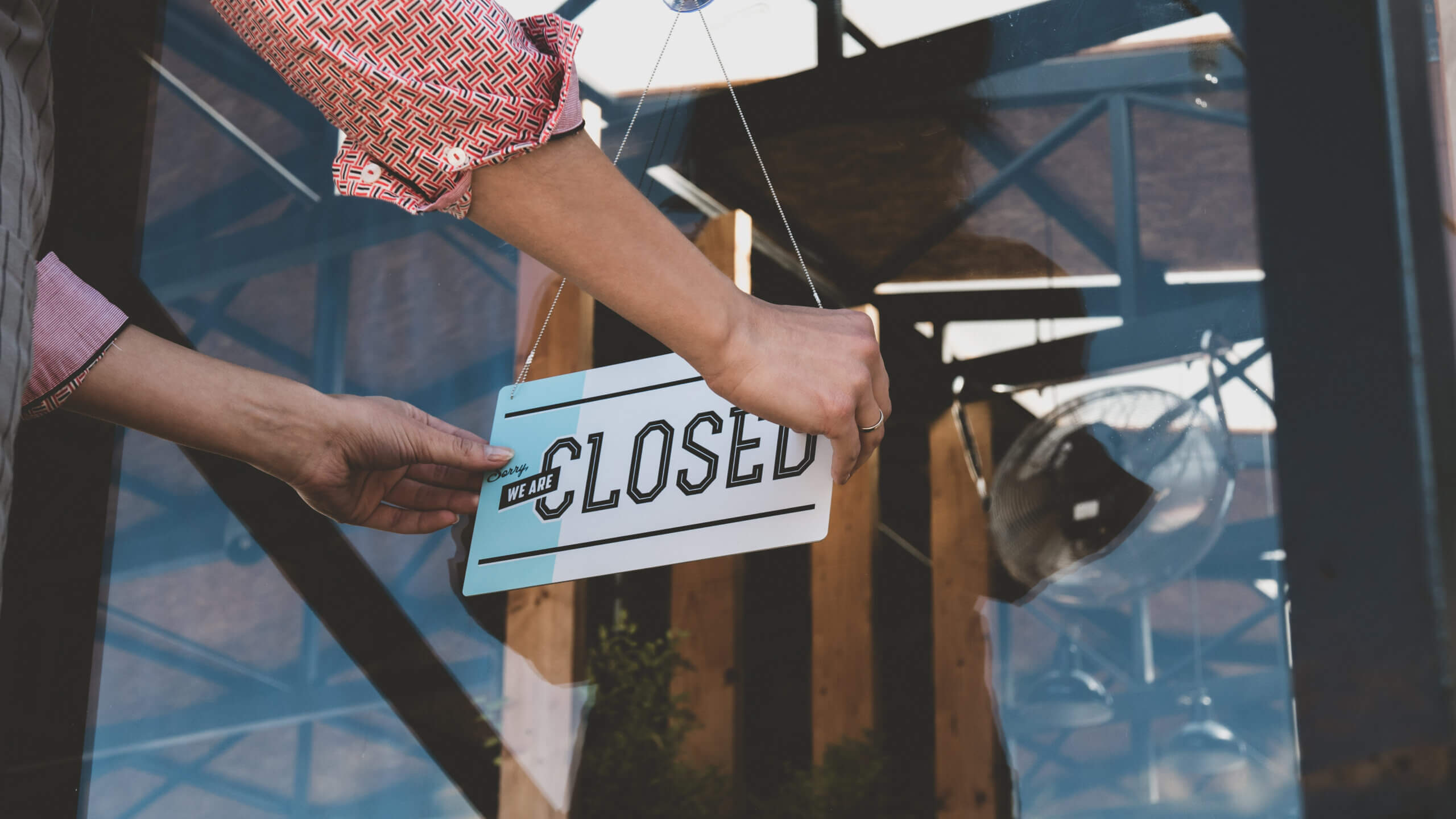 A person is hanging a "Closed" sign on the glass door of a shop or cafe. The scene reflects a sunny day with the structure of the building visible in the reflection. The person's sleeves are rolled up, showcasing a patterned shirt.
