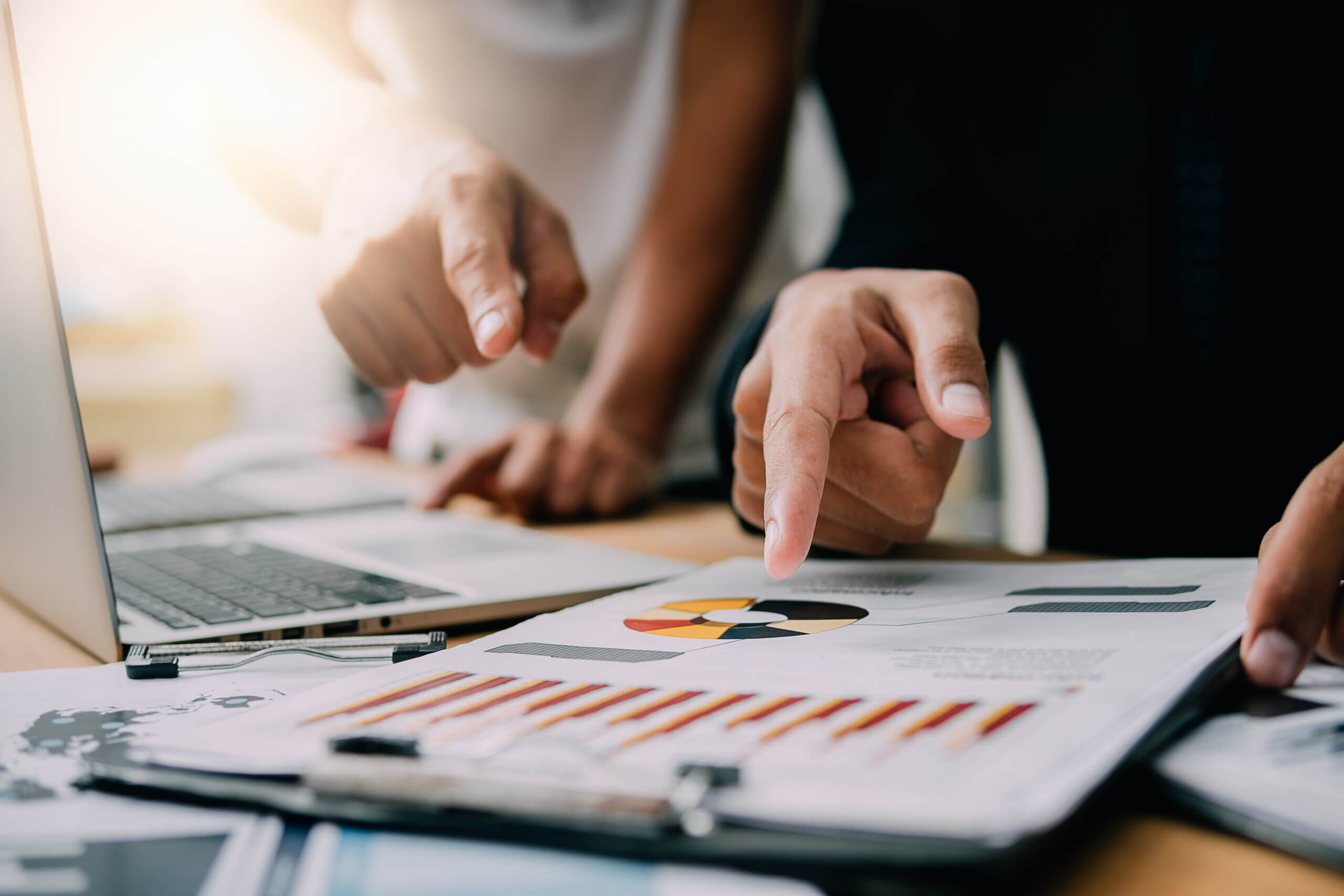 Close-up of two people pointing at graphs and charts on paper and a laptop screen in an office setting. The charts include a pie chart and a bar graph, suggesting a discussion or analysis of data.