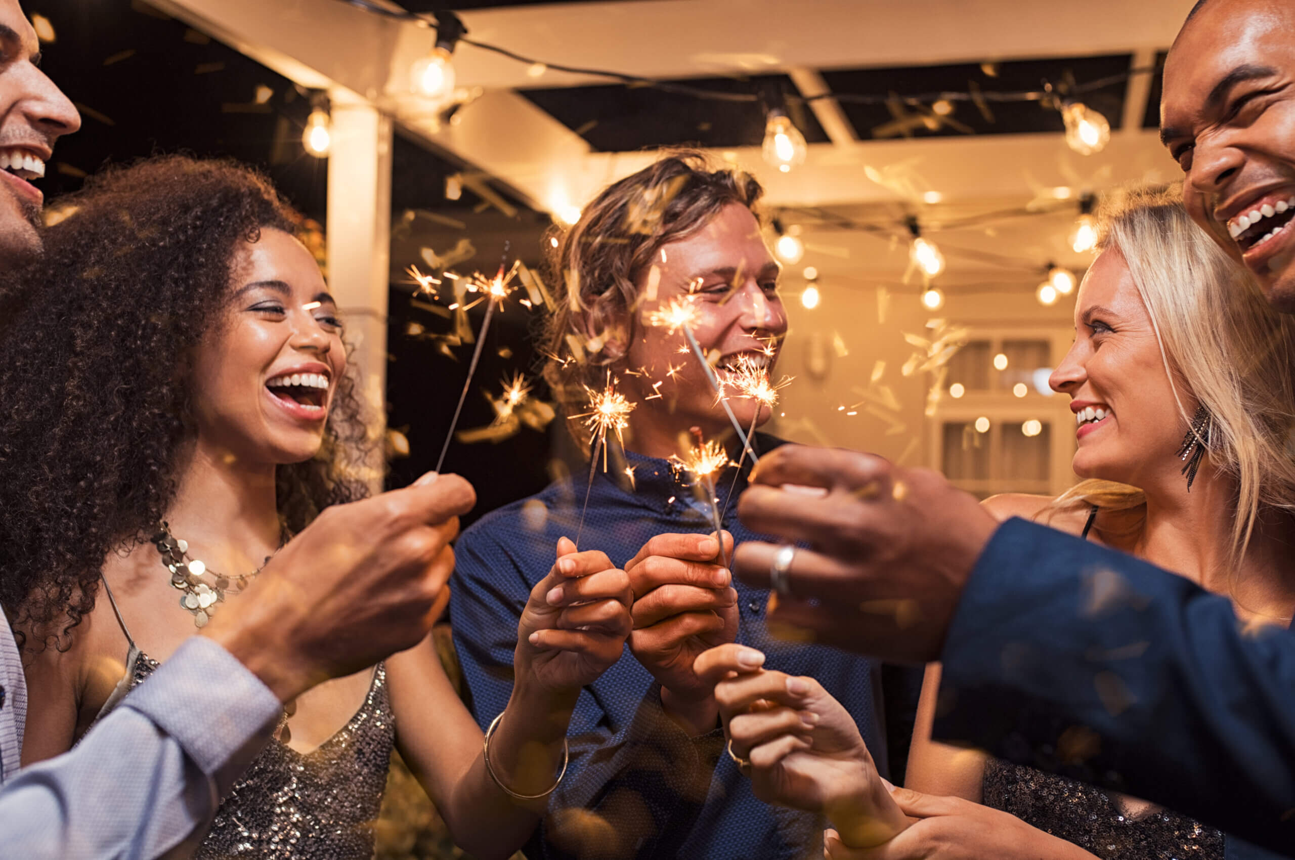 A group of people joyfully celebrating with sparklers in their hands. They are outdoors at night, surrounded by string lights, smiling and laughing. The atmosphere is festive and warm.