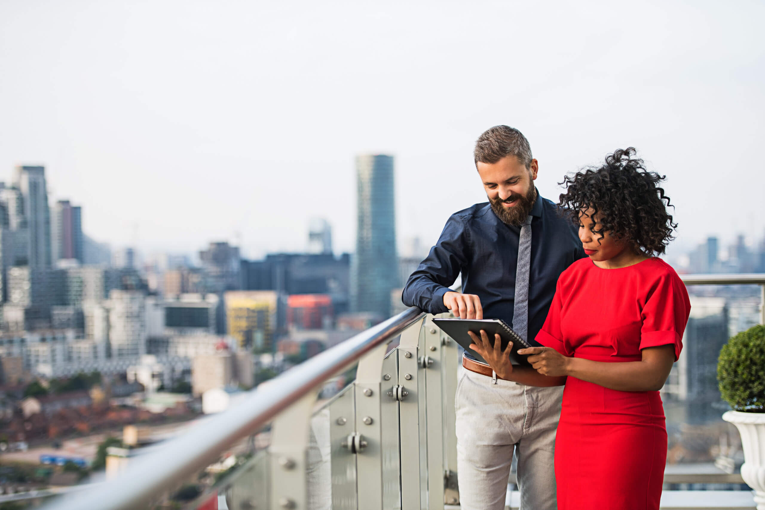 A man and a woman stand on a rooftop terrace overlooking a cityscape. The man wears a dark shirt and light trousers and points at a tablet held by the woman, who is wearing a red dress. Tall buildings are visible in the background.