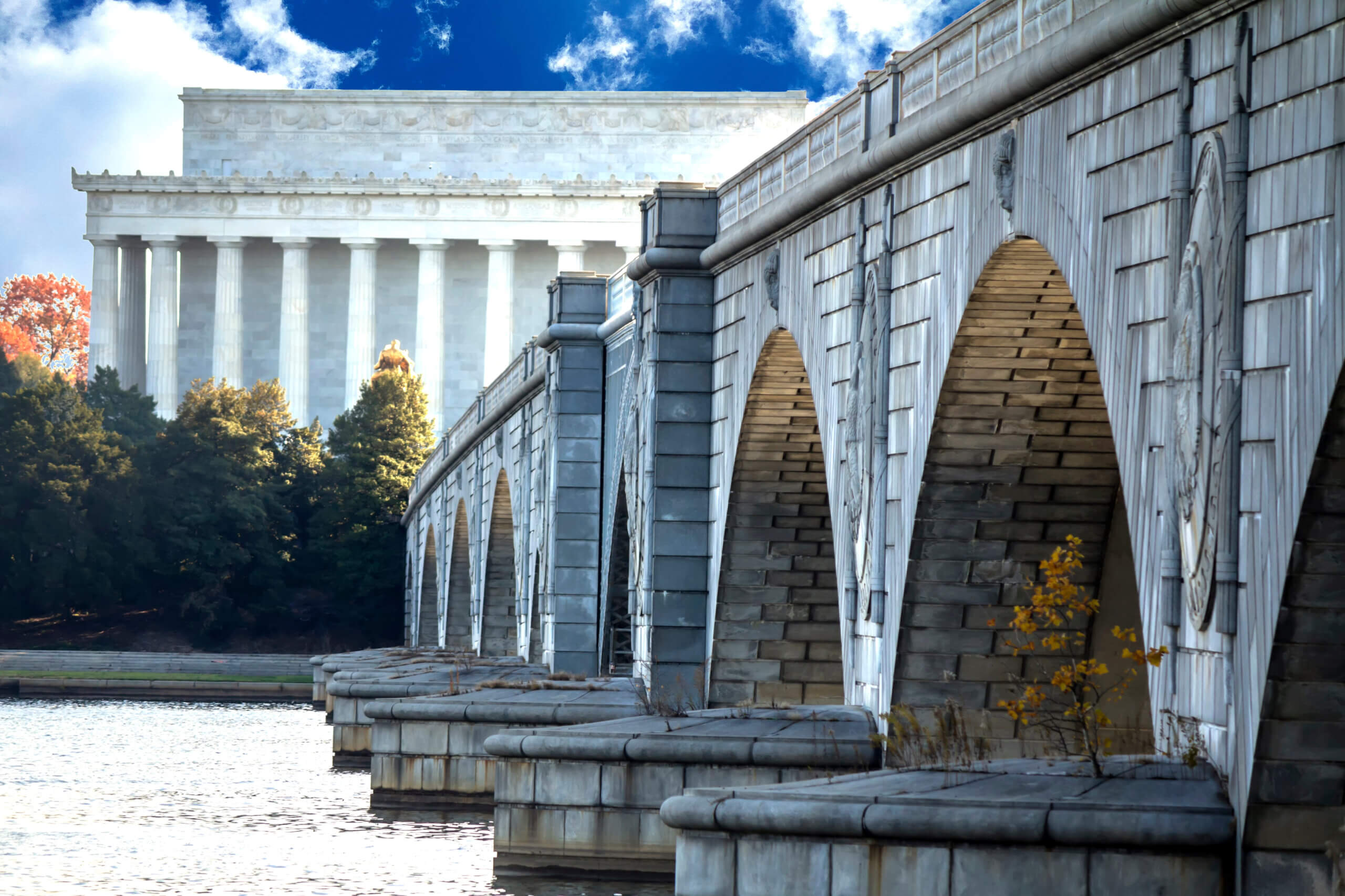 The image shows the Arlington Memorial Bridge spanning a river, leading to the Lincoln Memorial in the background. The bridge's stone arches reflect in the water, and trees with autumn leaves are visible nearby under a partly cloudy sky.