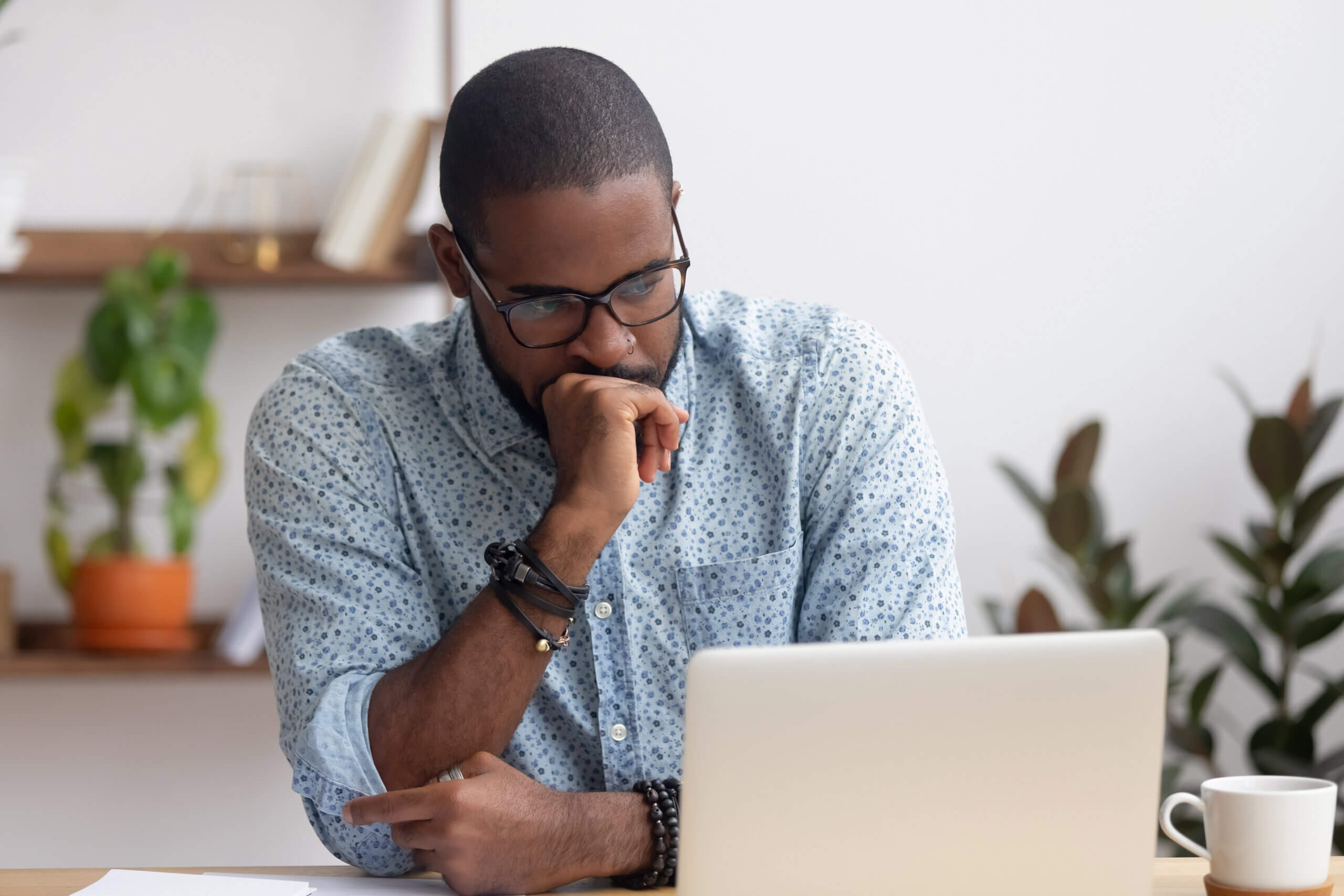 A person wearing glasses and a patterned shirt sits thoughtfully at a desk, looking at a laptop. A potted plant and a coffee cup are in the background, adding a touch of greenery and a casual atmosphere to the scene.