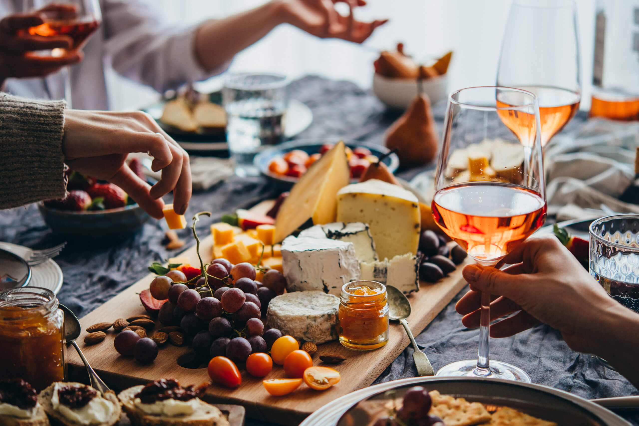 A group of people enjoying a charcuterie board with various cheeses, grapes, nuts, and cherry tomatoes. Two glasses of rosé wine are in the foreground, and hands are reaching for food items. The scene is set on a textured tablecloth.
