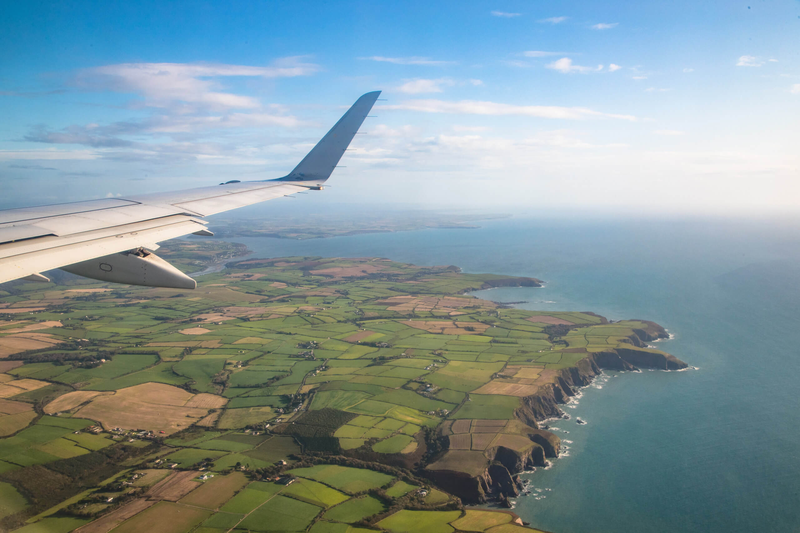 View from an airplane window showing a wing and a scenic landscape below. The land is divided into patchwork fields alongside a rugged coastline, with blue ocean and sky in the background.