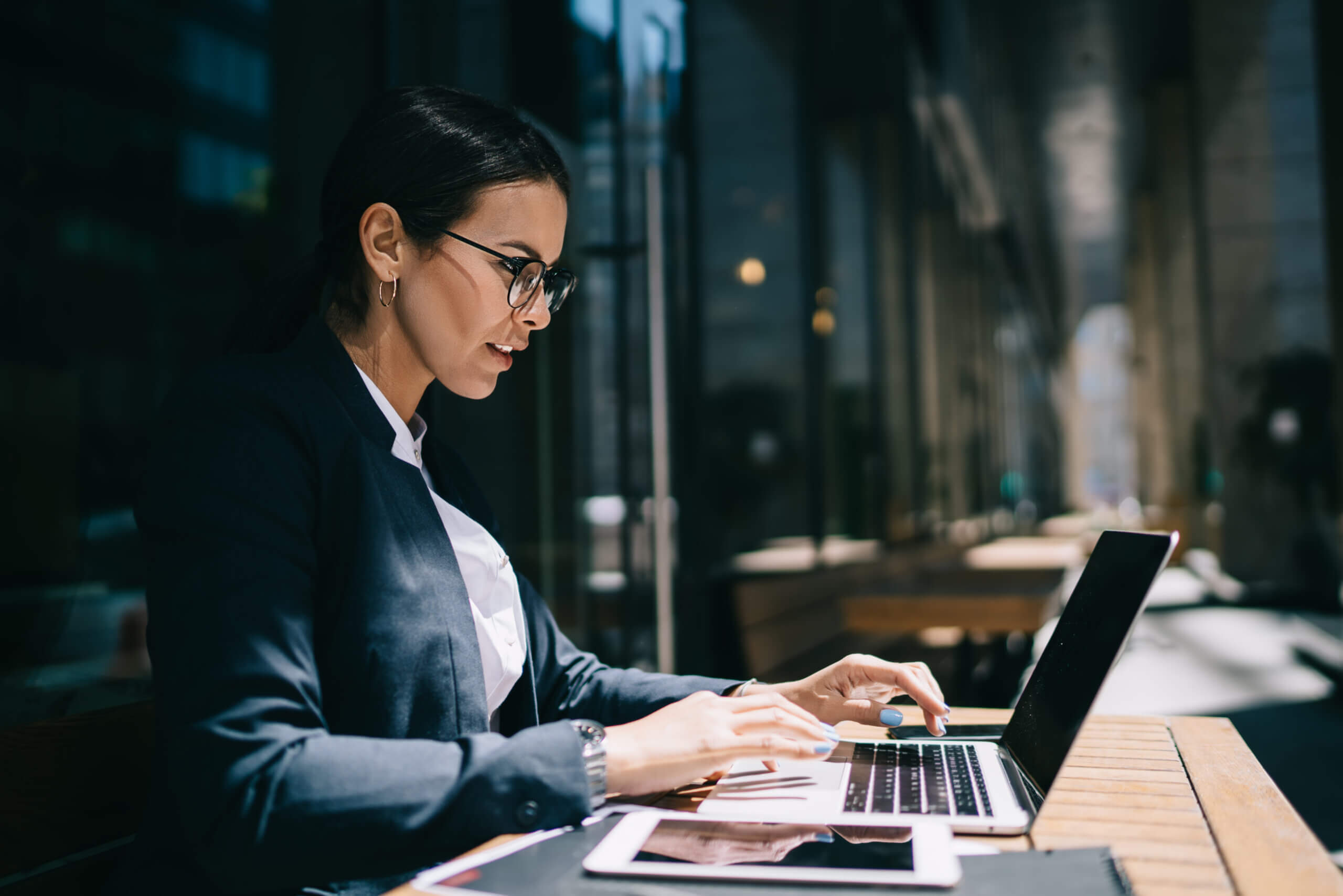 A person wearing glasses and a dark blazer works on a laptop outdoors at a wooden table. They are focused, and a tablet and phone are on the table beside them. The background shows a city street with tall buildings.