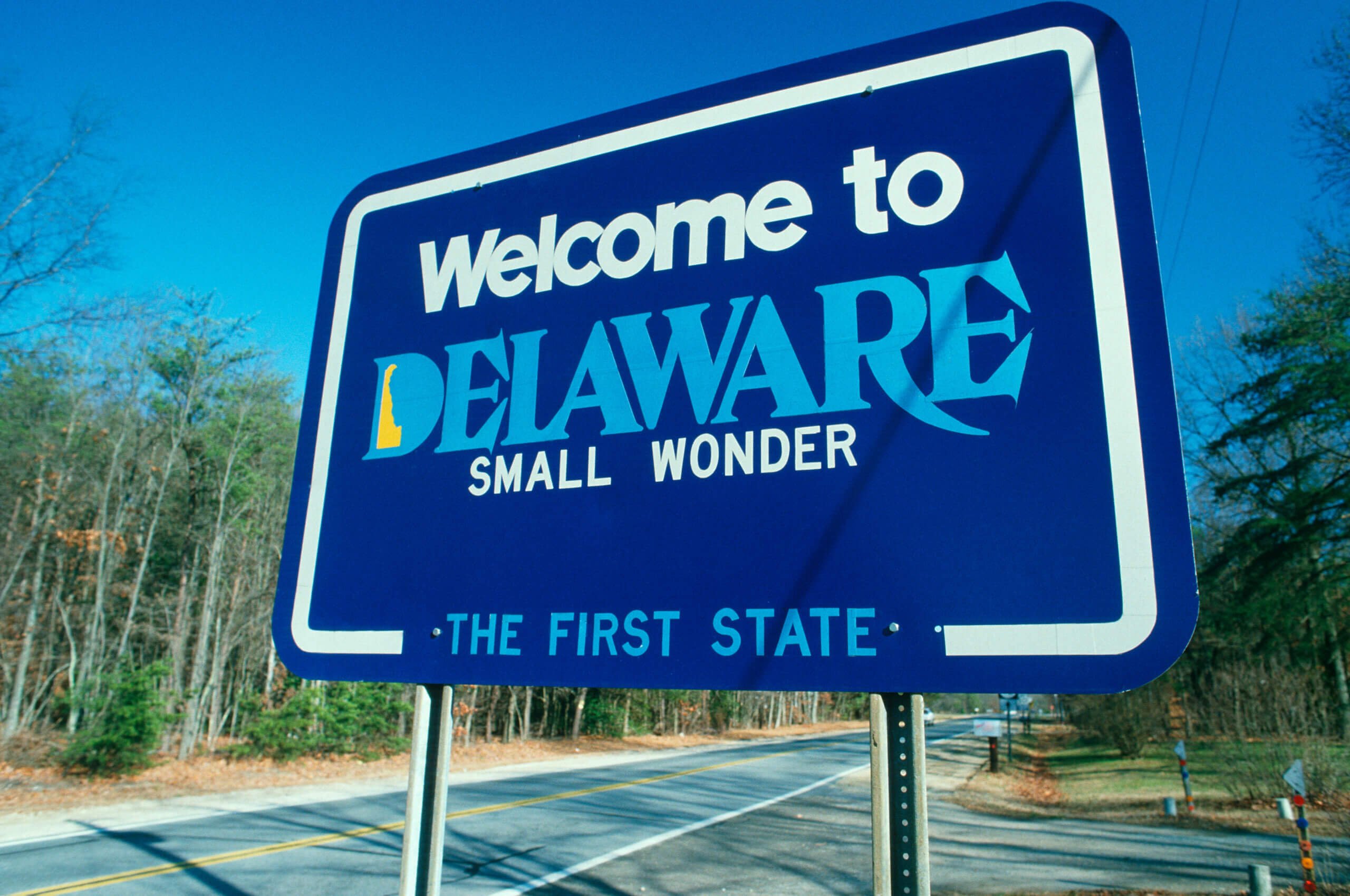 A blue roadside sign reads "Welcome to Delaware, Small Wonder, The First State." It is set against a backdrop of a road and trees with clear skies.