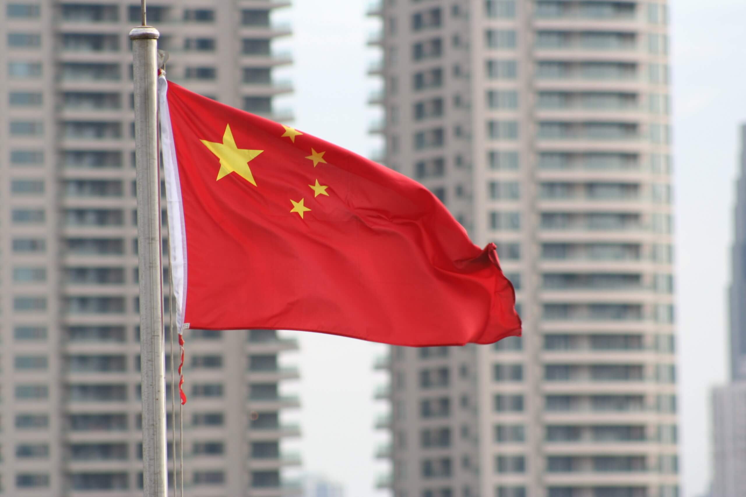 The Chinese flag waves on a pole in the foreground, featuring one large yellow star and four smaller stars on a red background. Blurred tall buildings are visible in the background.
