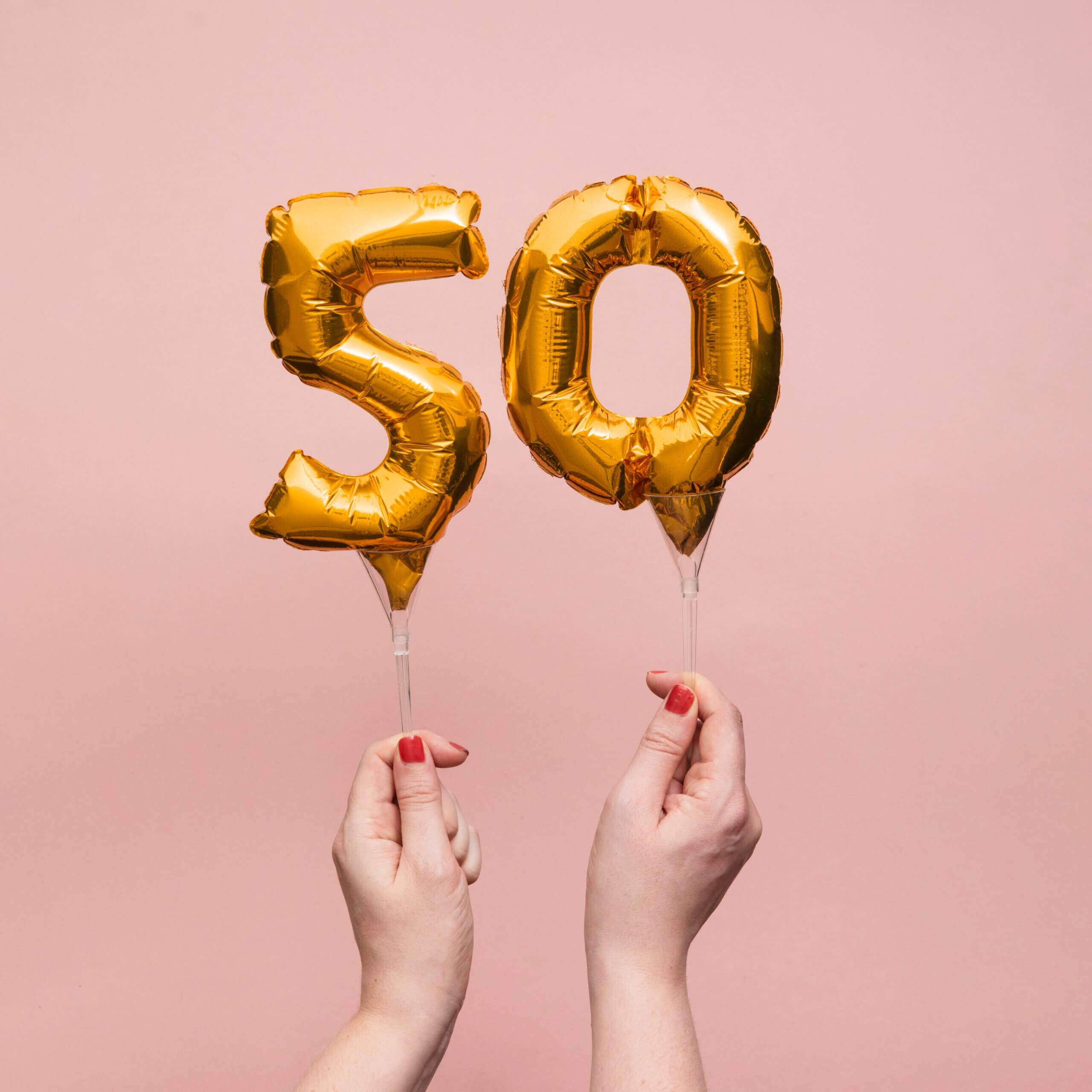 Hands holding gold foil balloons shaped like the number 50 against a pink background.