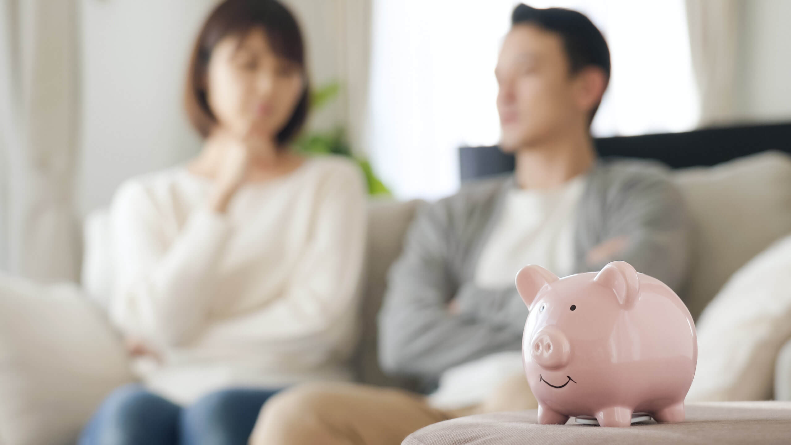 A pink piggy bank is in focus on a table, with a man and woman sitting on a couch in the background, appearing blurred. They seem contemplative, with the woman resting her chin on her hand. The setting is indoors.