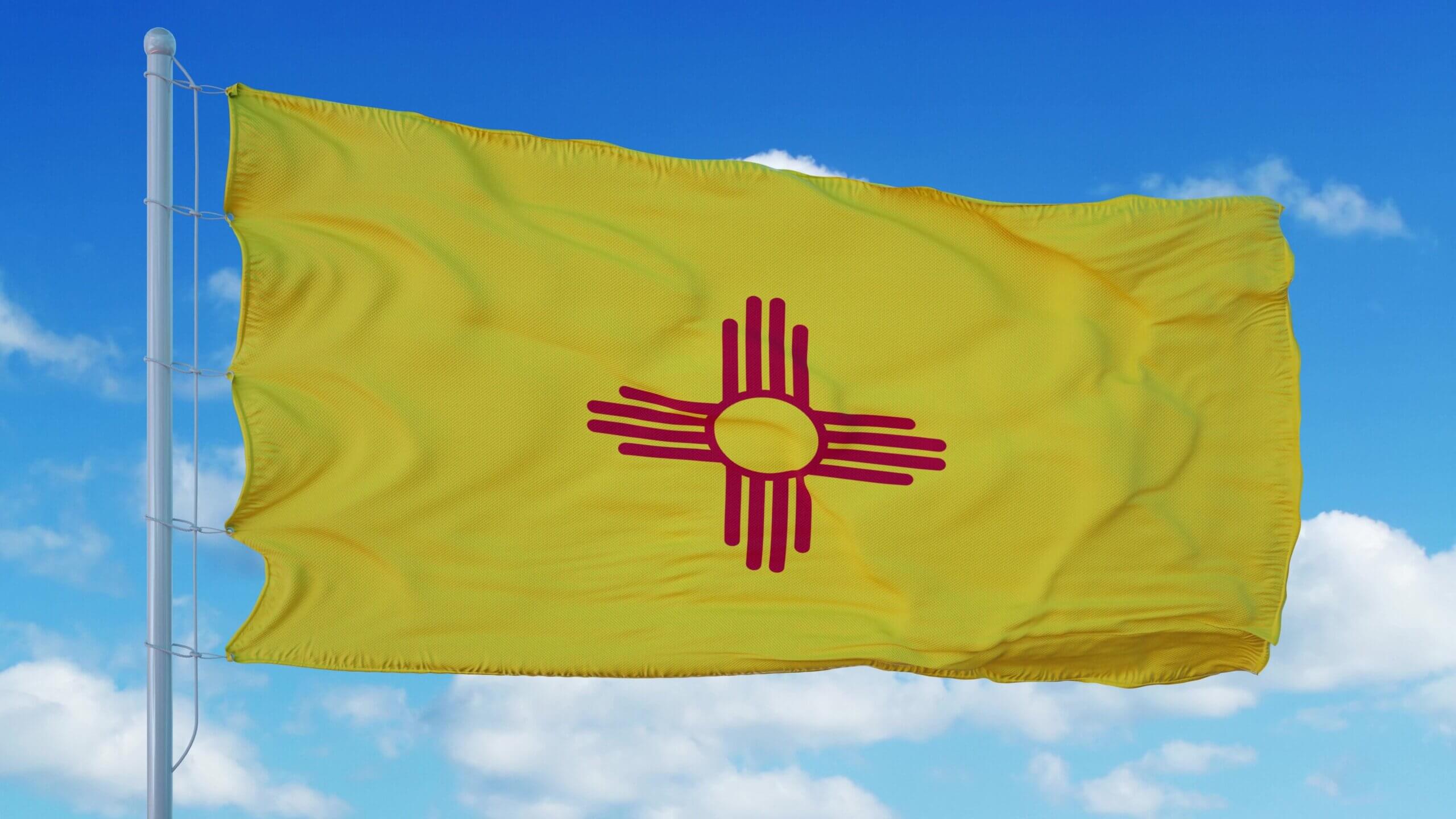 The image shows the New Mexico state flag, featuring a yellow background with a red Zia sun symbol in the center, blowing in the wind against a blue sky with scattered clouds.