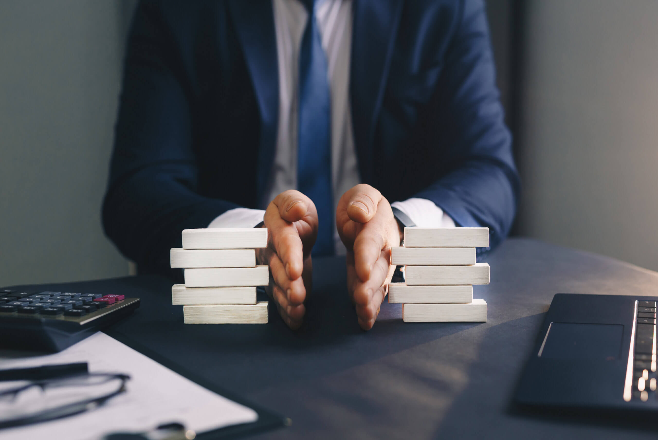 A person in a suit is seated at a desk, using both hands to separate stacks of white wooden blocks into two groups. Nearby are a calculator, some papers, and a laptop, suggesting a business or financial setting.