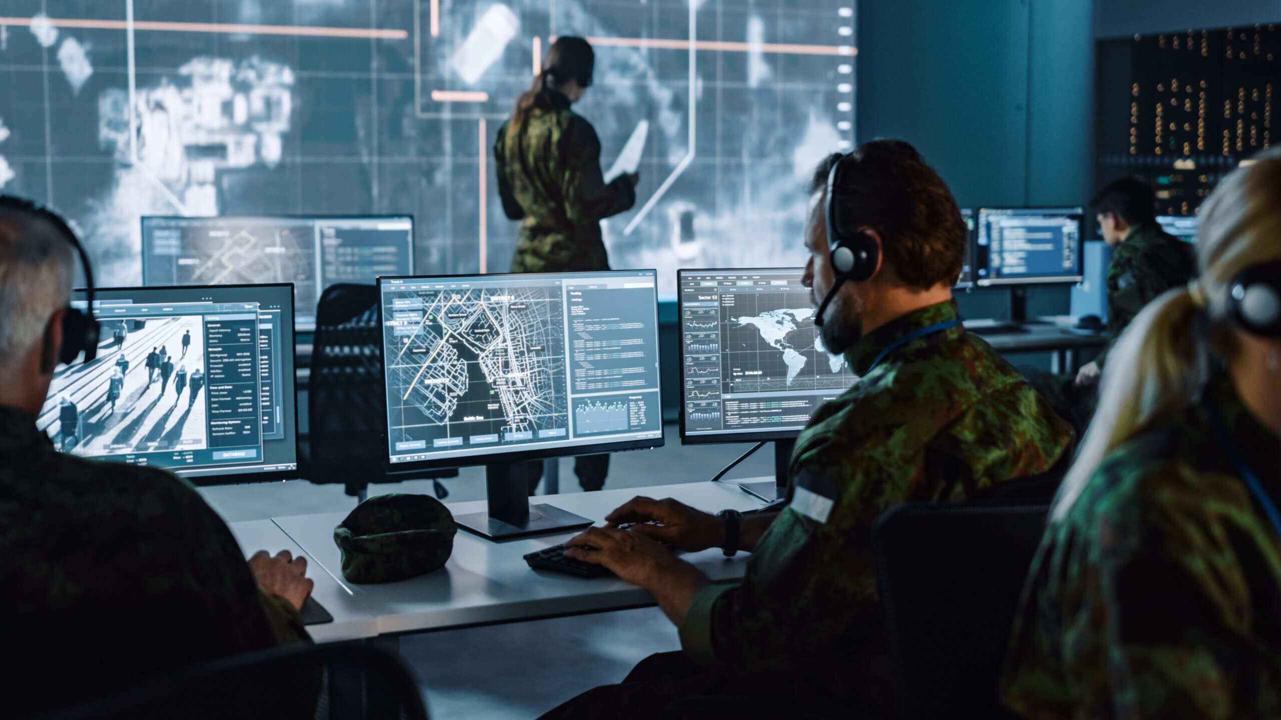 A group of military personnel work at computer stations in a dim control room, analyzing maps and data on multiple monitors. A large digital map is projected on a screen in the background. They wear camouflage uniforms and headsets.