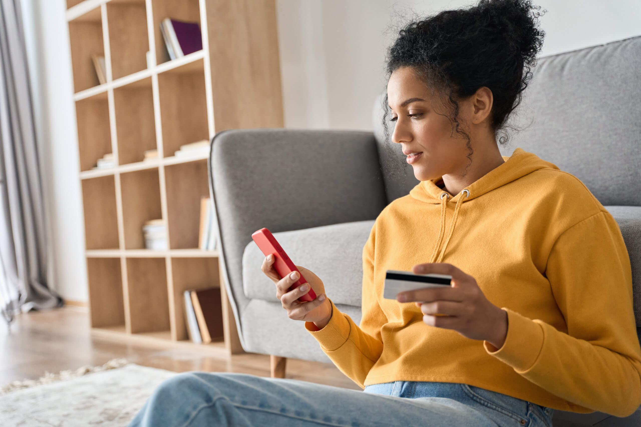 A woman in a yellow hoodie sits on the floor holding a smartphone and a credit card. She appears focused, possibly making an online purchase. In the background, there's a gray sofa and a wooden bookshelf with books.