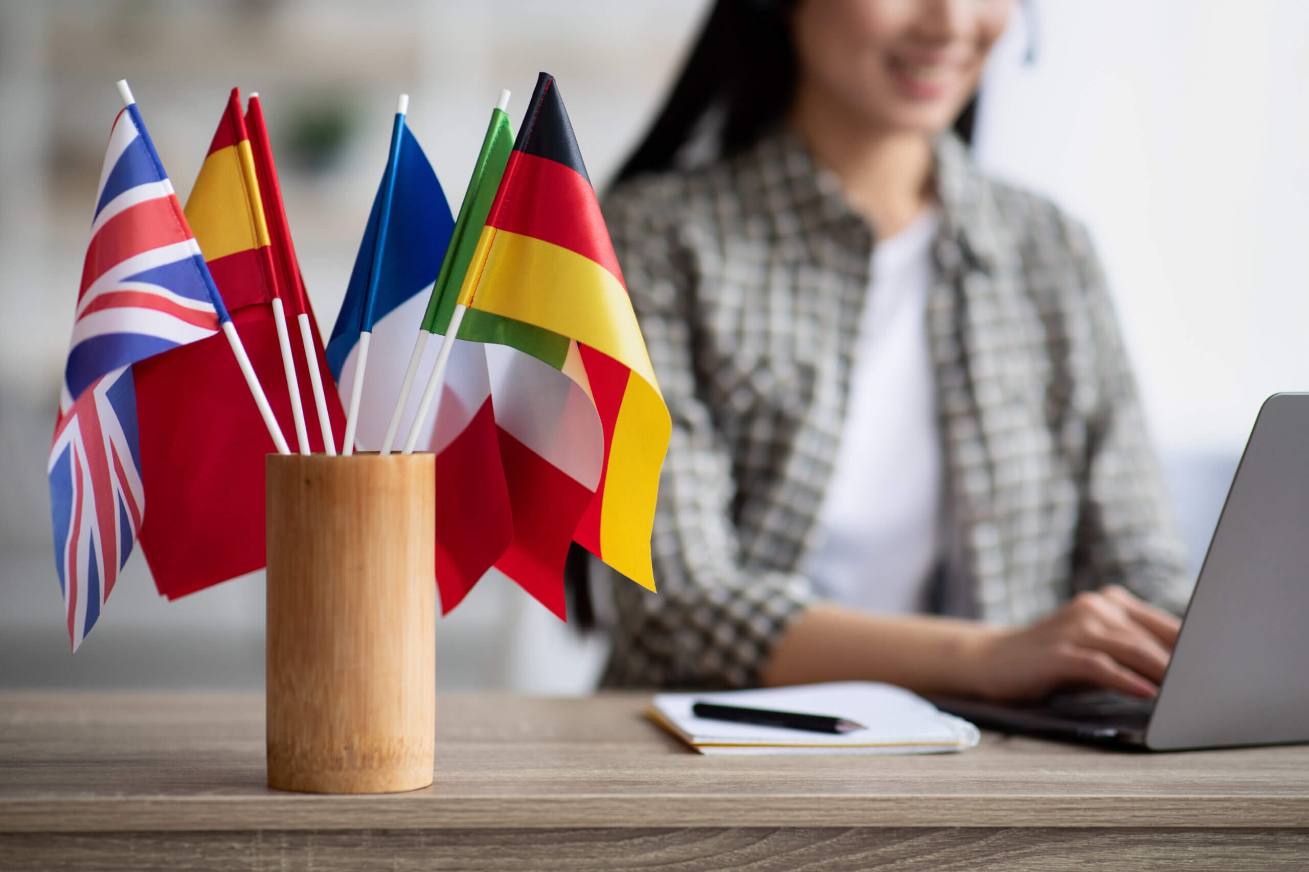 A wooden holder contains small flags of different countries, including the UK, Spain, France, Germany, and Italy. In the background, a person wearing a plaid shirt is typing on a laptop, with a notebook nearby.