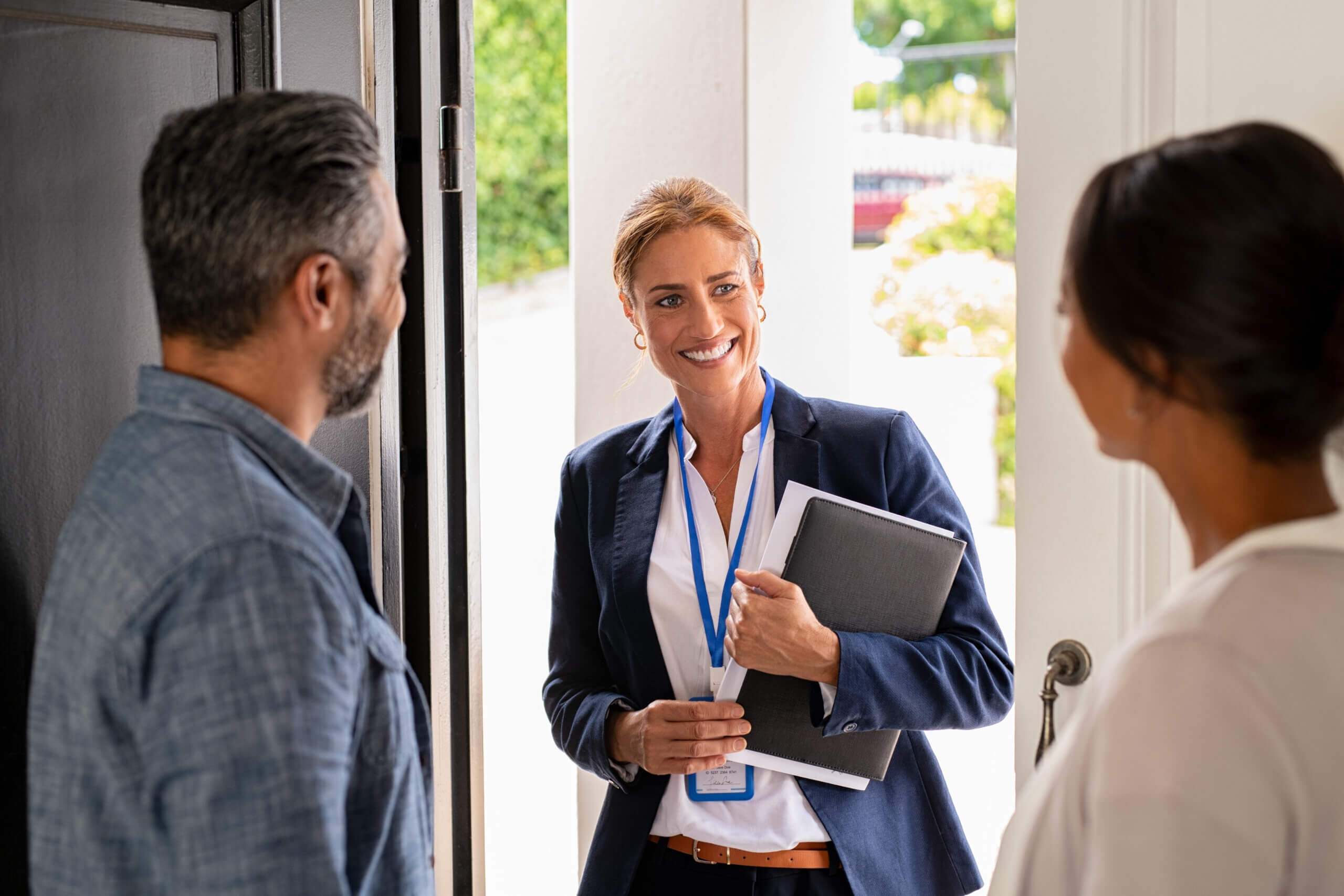 A woman in a blue blazer stands at a doorway, holding a notebook and smiling at a man and woman inside the house. The man has gray hair and a beard, wearing a denim shirt; the woman inside has long dark hair. Bright, sunny background visible outside.