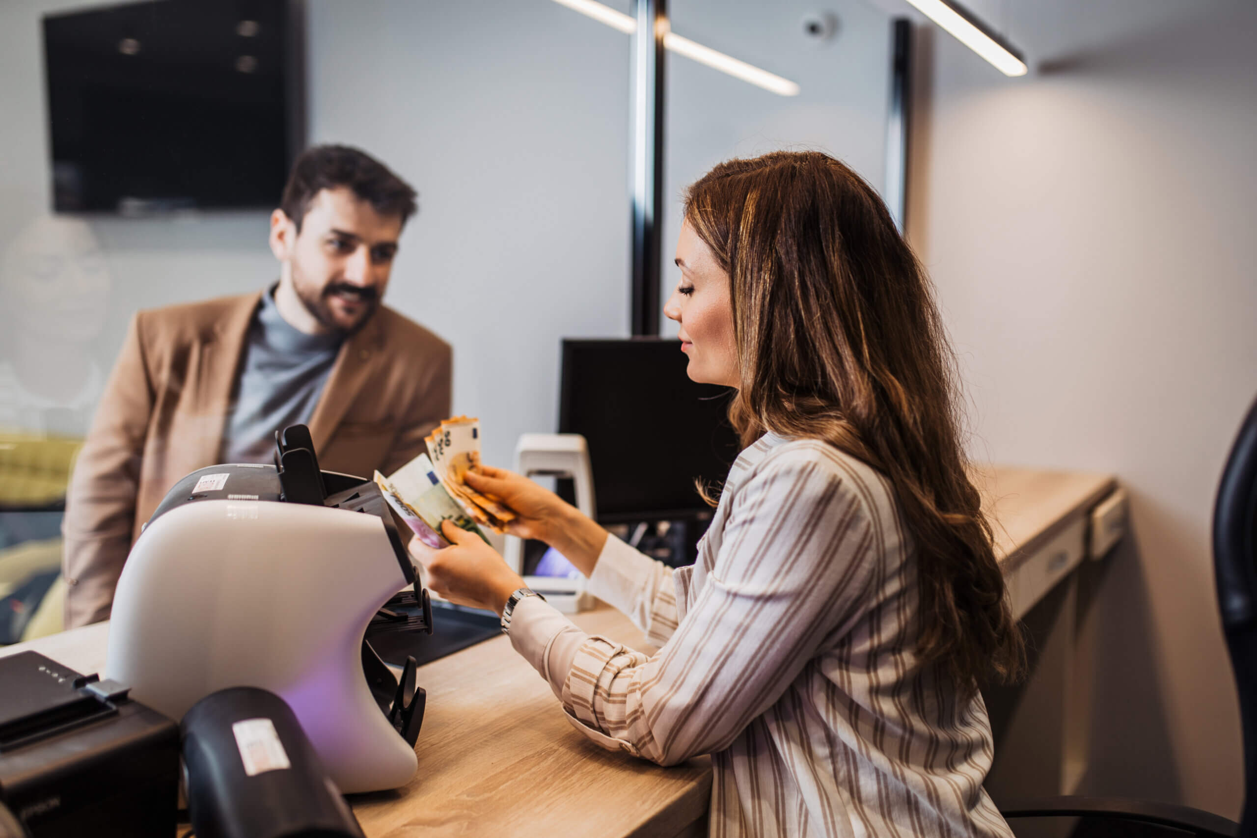 A woman in a striped shirt is sitting at a desk, handling some money, while a man in a brown jacket stands on the other side. There's a card machine and computer on the desk, and a TV mounted on the wall in the background.