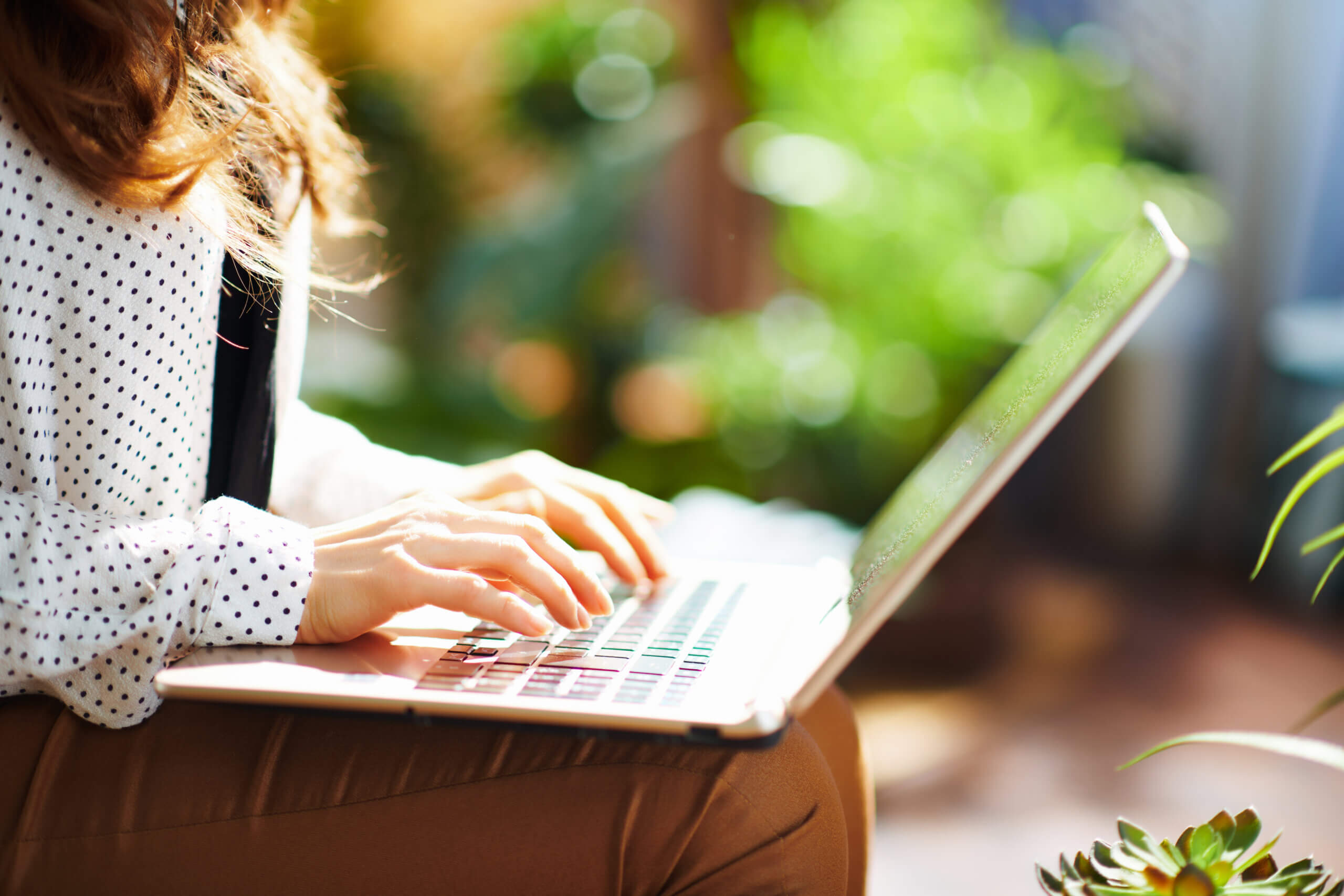 A person in a white polka dot shirt types on a laptop while sitting outside. The background is filled with green plants, creating a bright and natural setting.