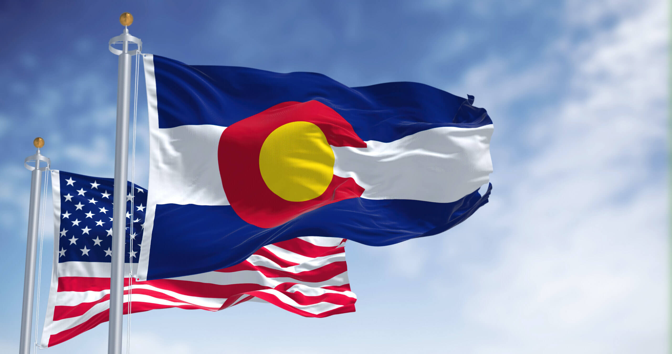 Two flags waving against a blue sky: the Colorado state flag, featuring blue, white, and red with a yellow circle; and the United States flag, with red and white stripes and a blue field of stars.