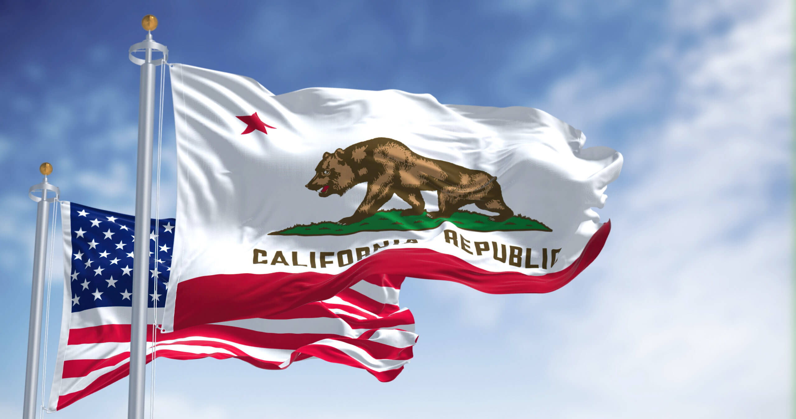 The image shows the California state flag and the United States flag waving in the wind against a blue sky. The California flag features a brown bear and a red star above "California Republic." The U.S. flag has red and white stripes and white stars on a blue field.