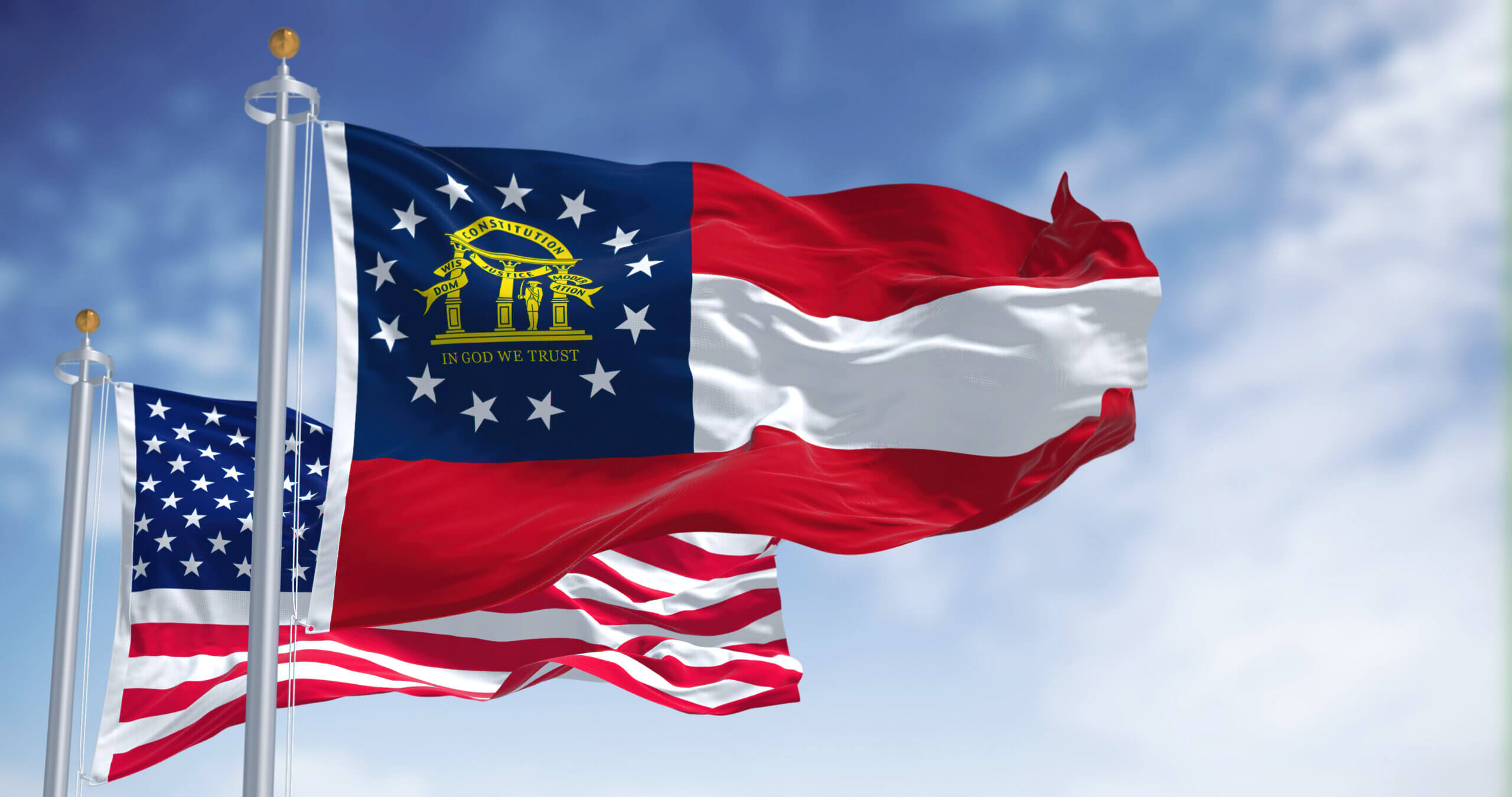 The image shows the Georgia state flag, with the motto "In God We Trust," flying in the foreground alongside a U.S. flag, both waving against a blue sky with scattered clouds.