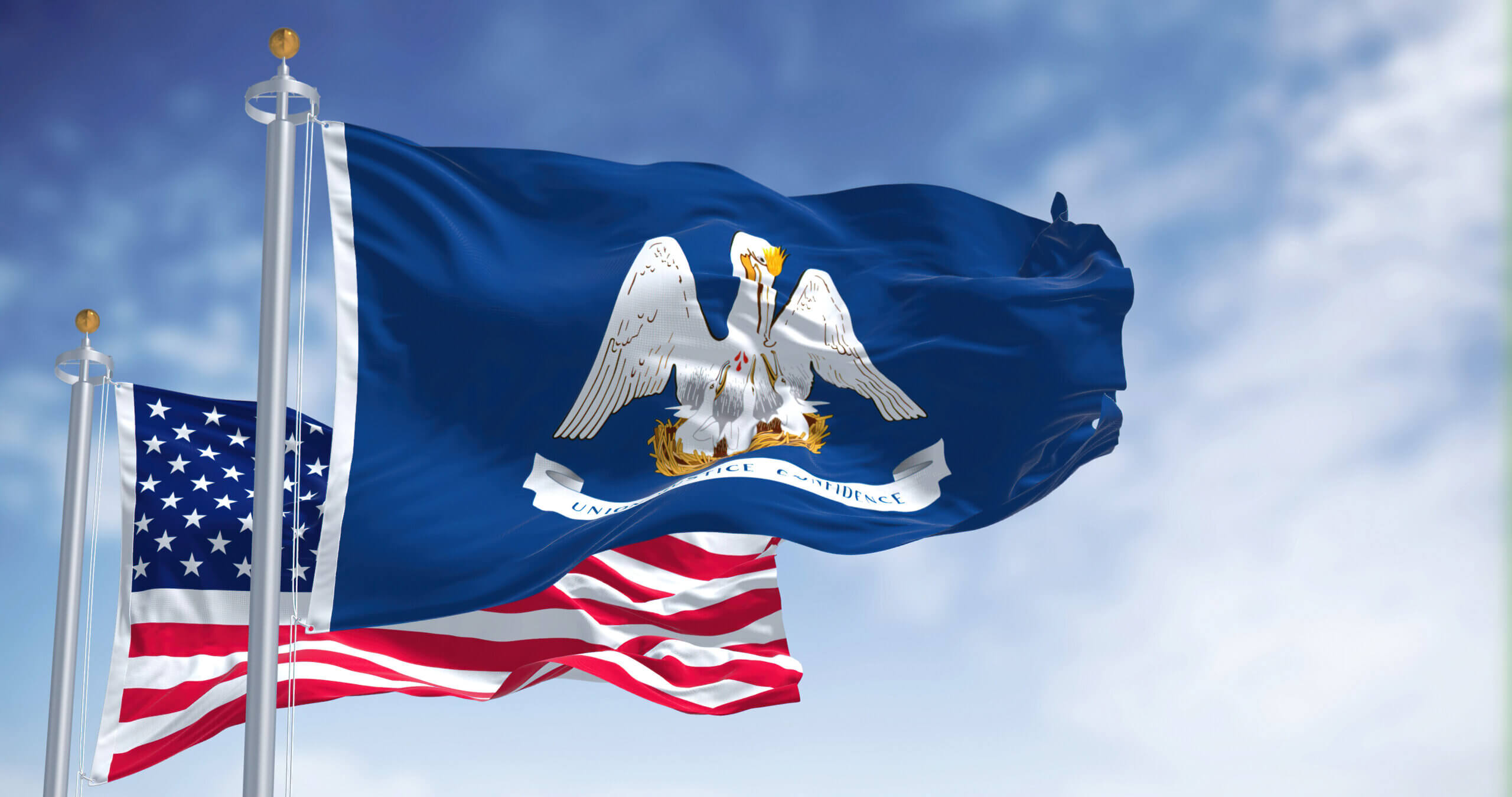 Two flags are flying against a blue sky with clouds. The flag in the foreground is the Louisiana state flag featuring a pelican. Behind it, partially visible, is the United States flag.