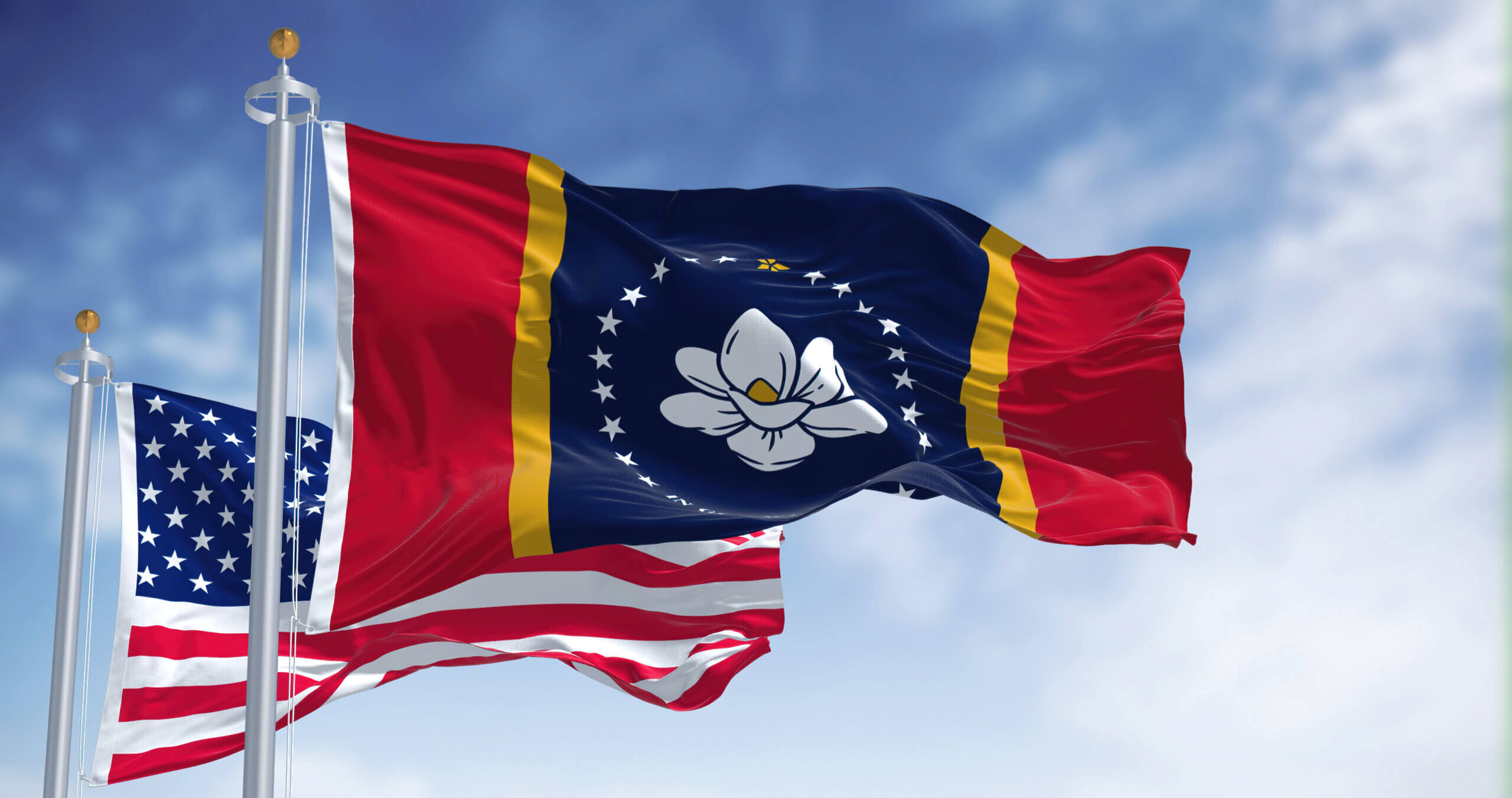 Two flags waving: one is the U.S. flag with stars and stripes, and the other features a blue field with a central magnolia flower, surrounded by stars within yellow and red borders, set against a partly cloudy sky.