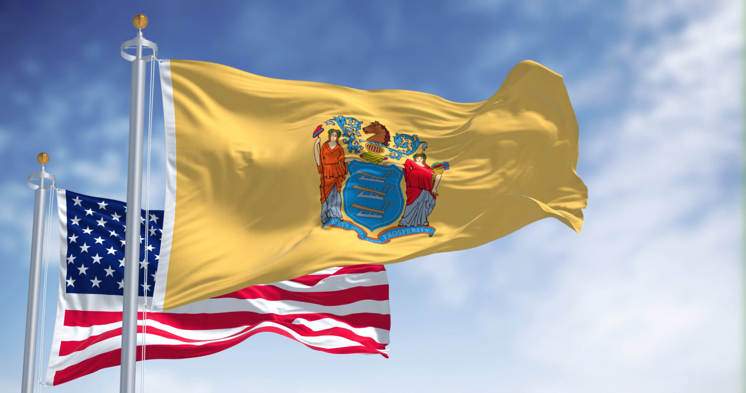 Flags of the United States, New Jersey, and an unidentified flag wave against a blue sky with clouds. The New Jersey flag is yellow with a blue shield and crest in the center. The U.S. flag is red, white, and blue with stars and stripes.