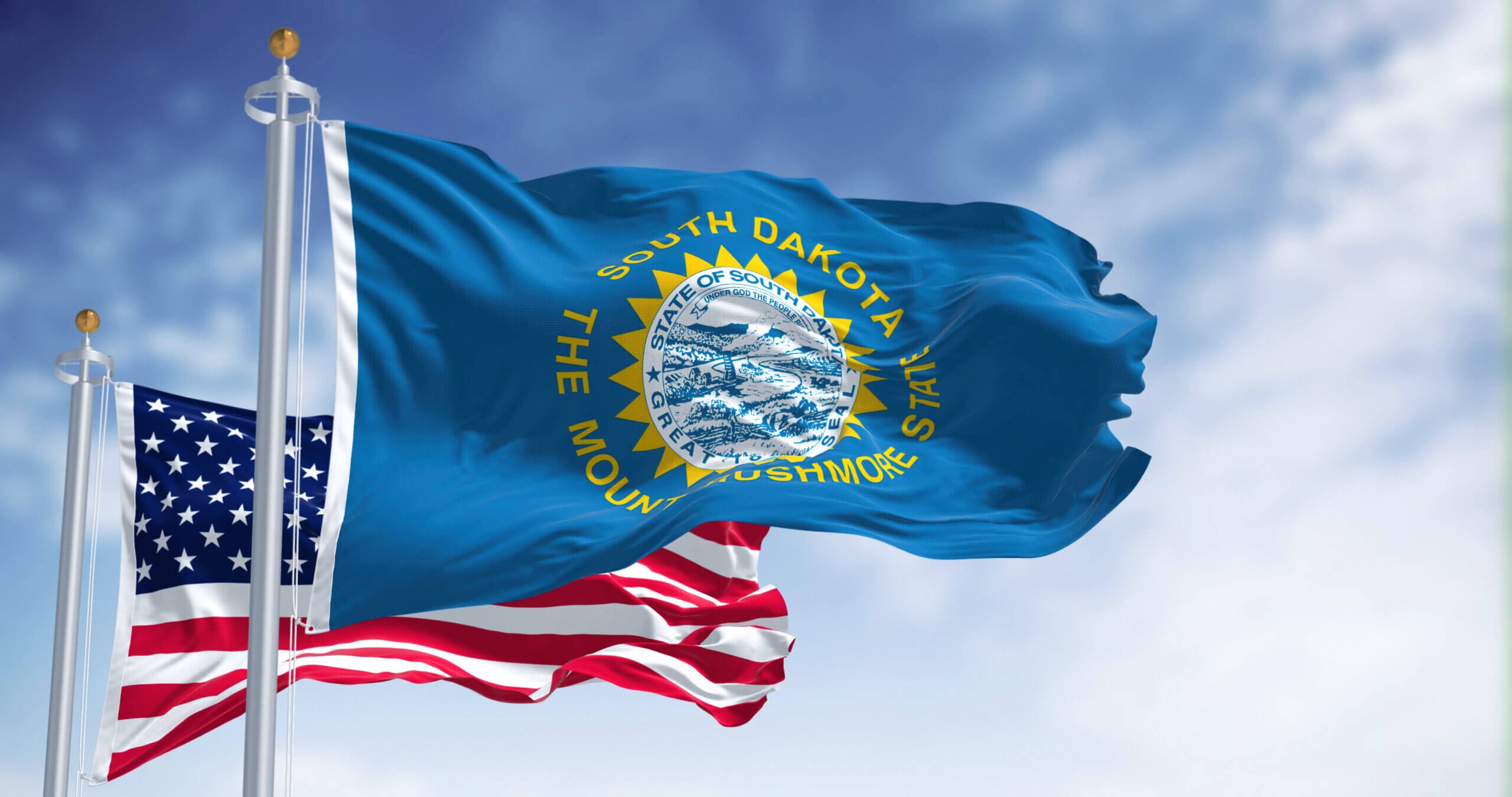 The image shows the South Dakota state flag and the American flag waving on flagpoles against a blue sky with clouds. The South Dakota flag features a state seal, sunburst design, and the text "South Dakota The Mount Rushmore State.